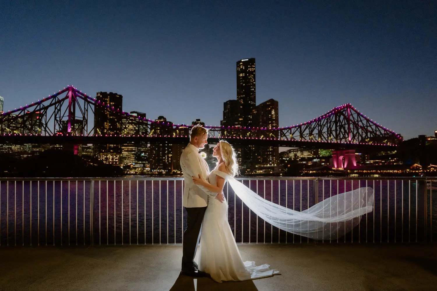 The couple at night on the river’s edge, the veil mid-air and the Story Bridge lit behind them, Bougainvillea House