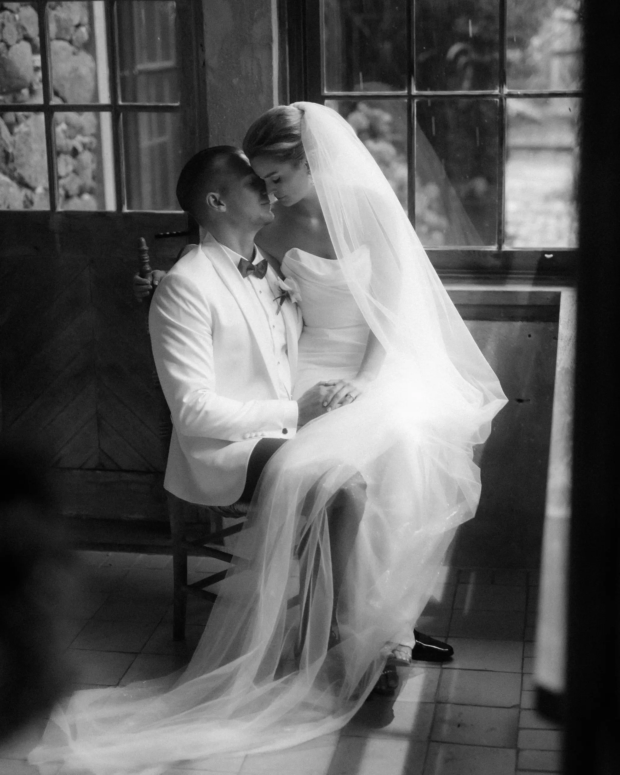 Bride sitting on the groom's lap by tall windows, her veil spread across the tiled floor
