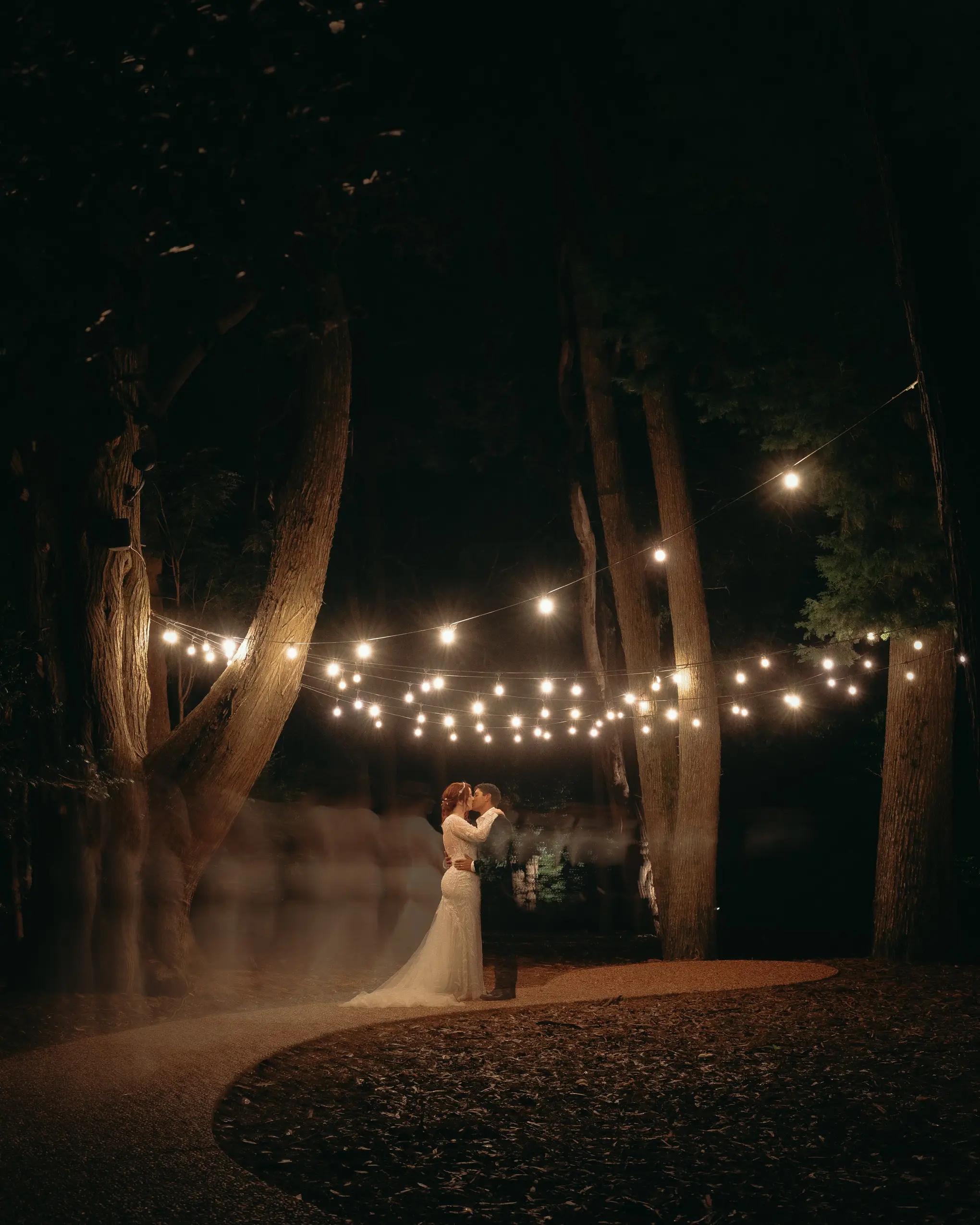 The couple kissing on the path after dark beneath strings of festoon lights, tall trees around them and guests blurred in the long exposure, Gabbinbar Homestead