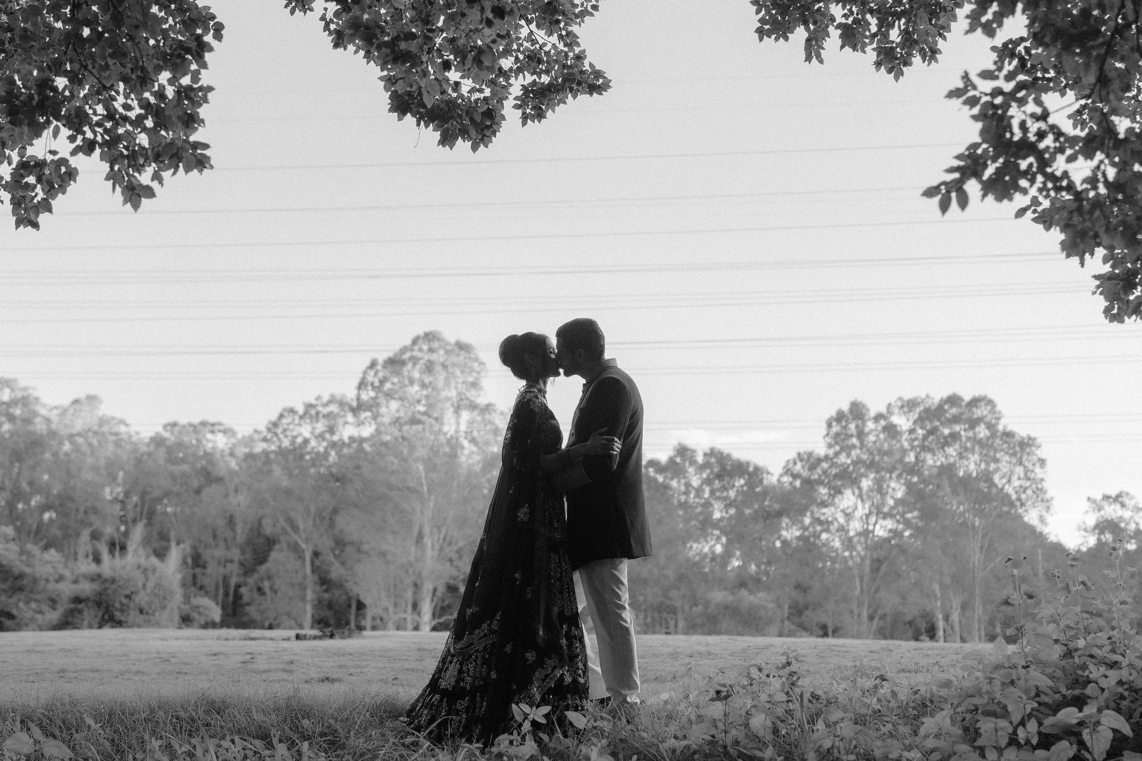Black and white photograph of a couple kissing in a paddock at dusk, the bride in an embroidered lehenga with tree branches framing the sky above