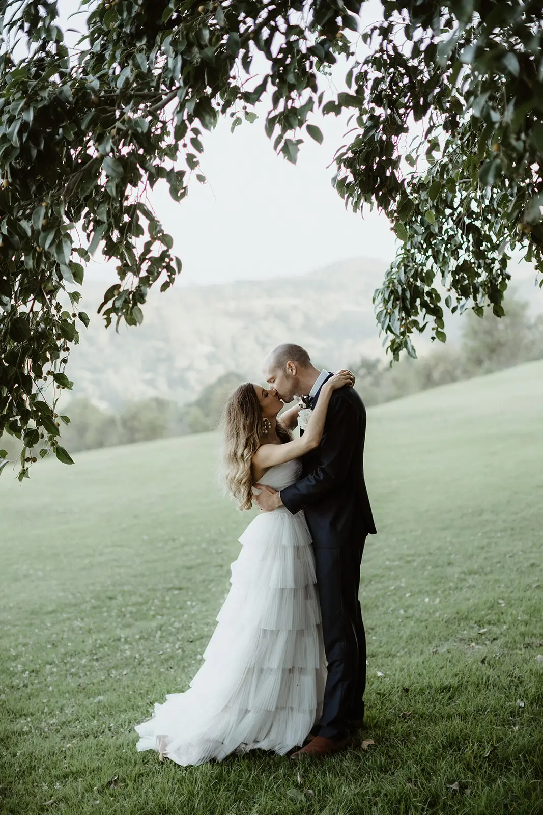 The couple close together under a tree on the open lawn, hills beyond, Summergrove Estate