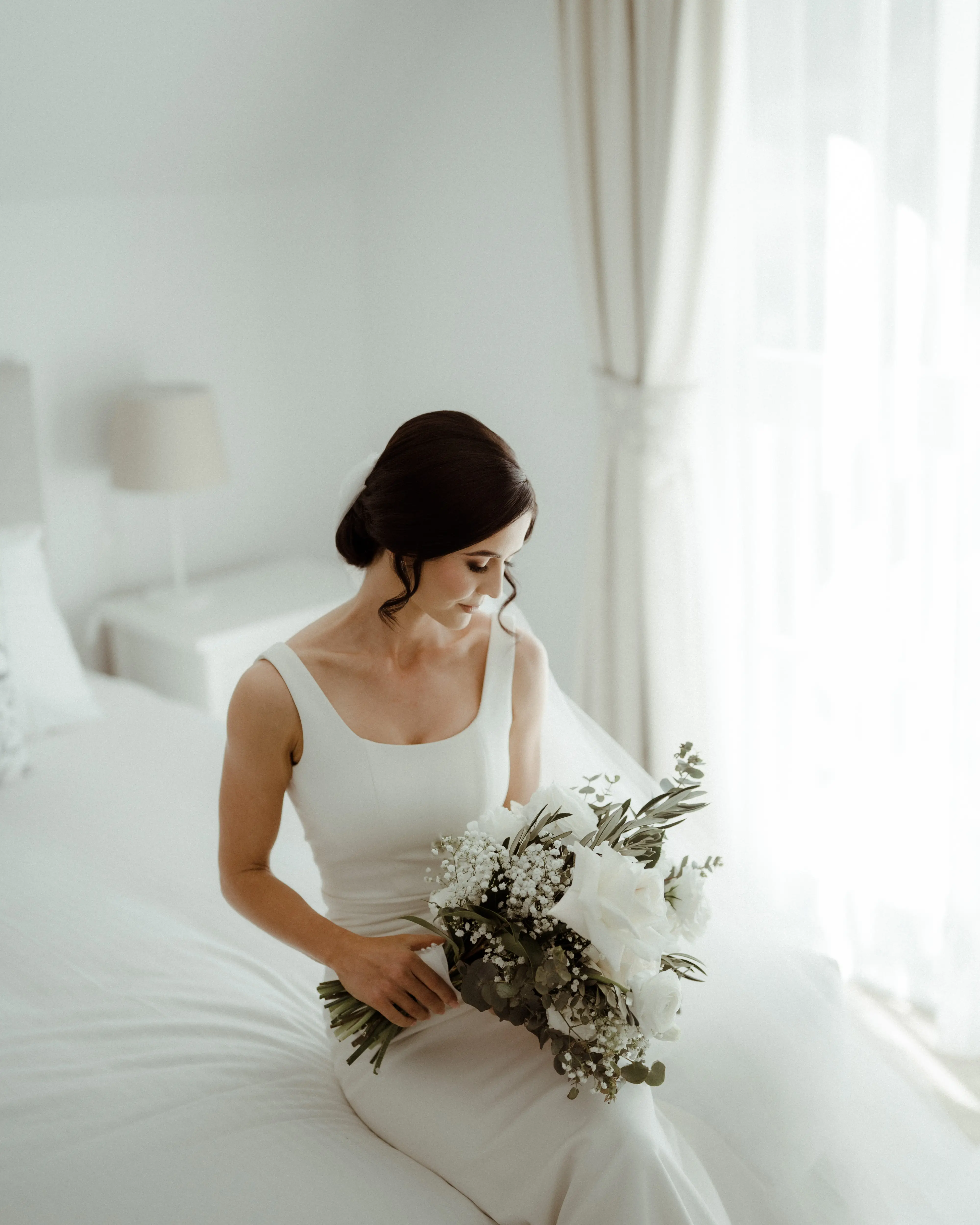Bride seated on the edge of a bed in soft window light, holding her bouquet