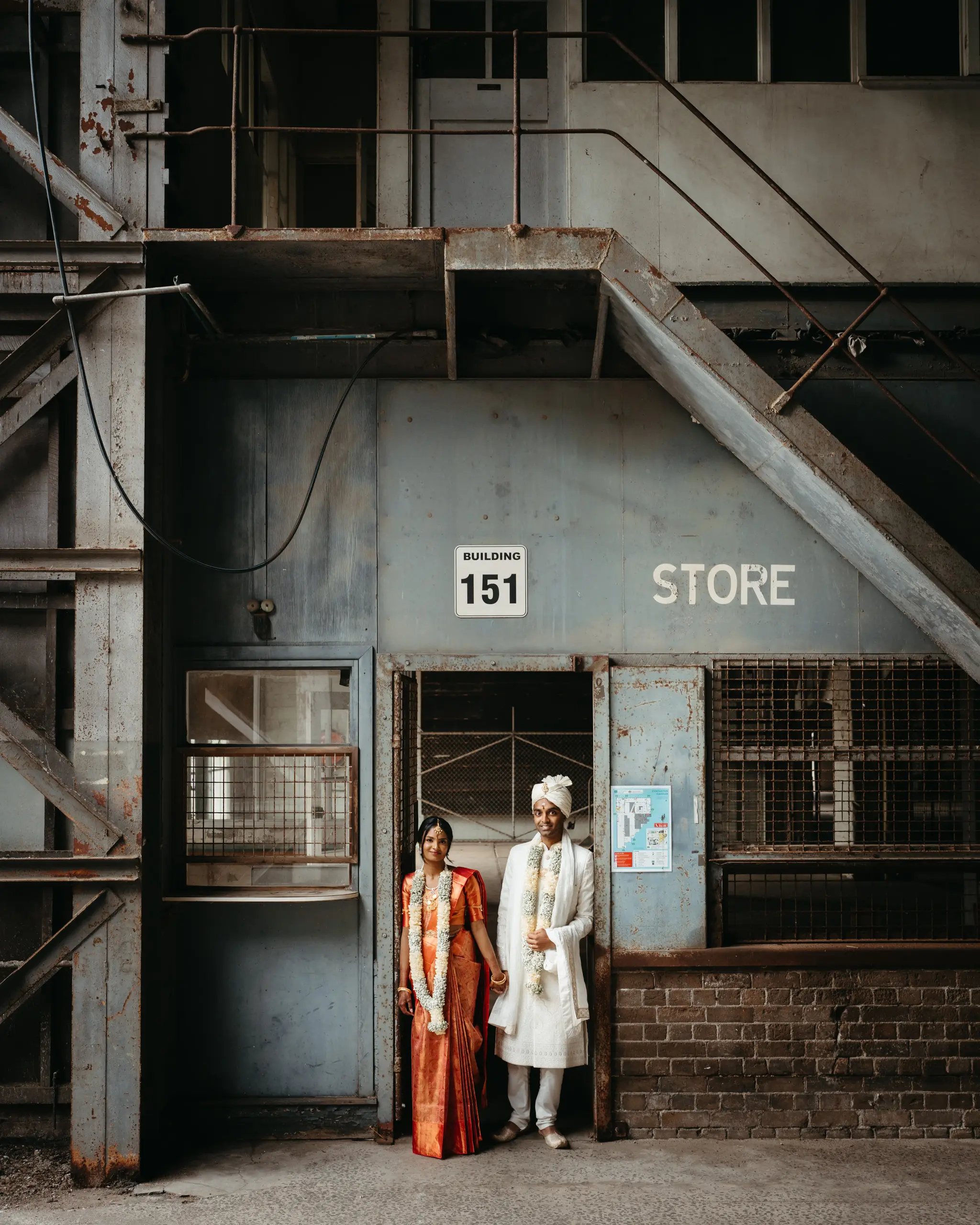 Couple in red sari and white sherwani outside a weathered industrial store
