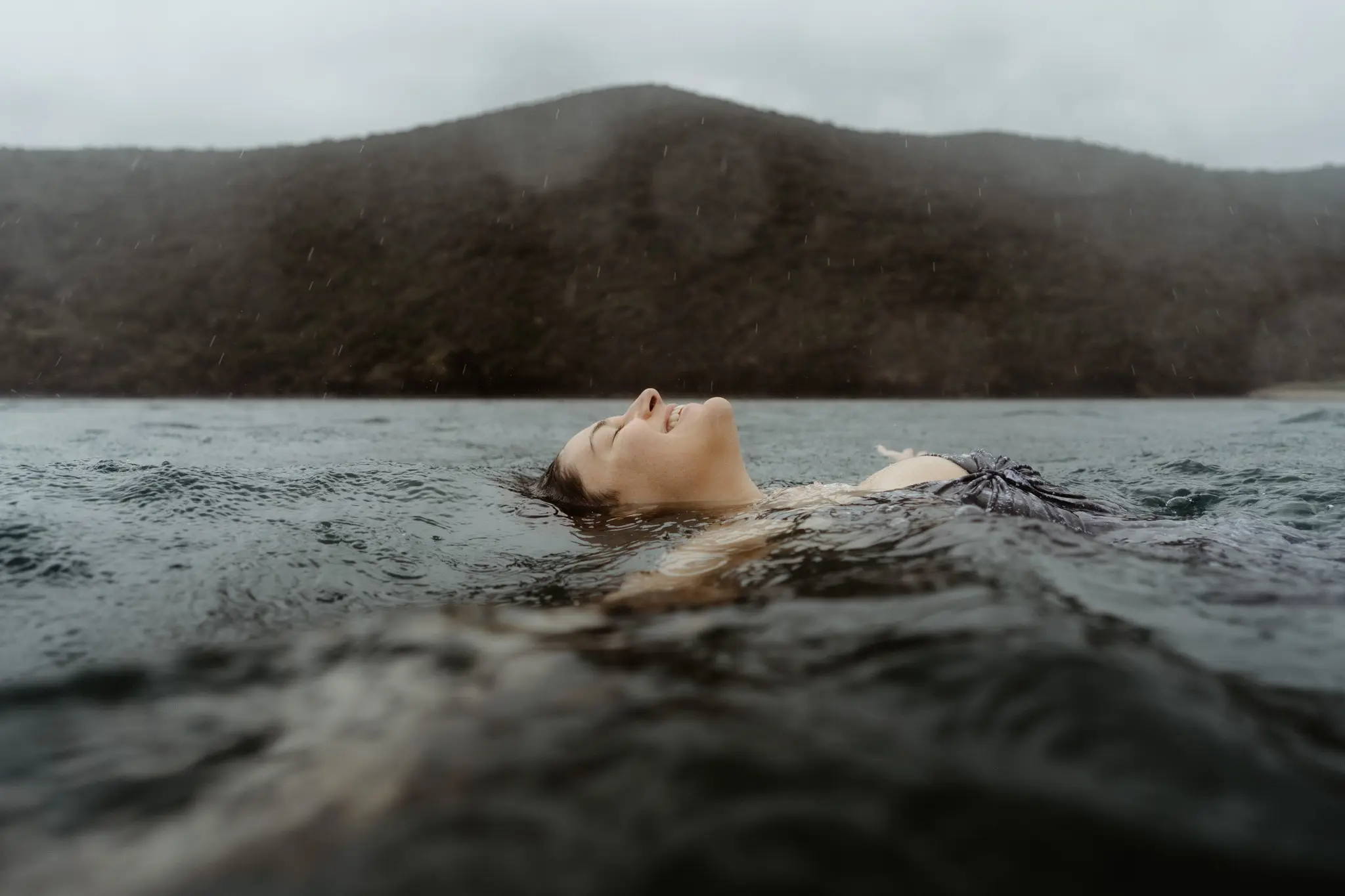 Bride floating on her back in dark water beneath a mountain ridge