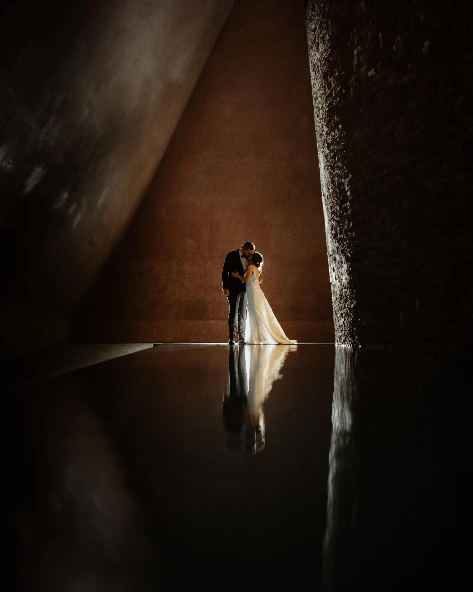 Bride and groom reflected at the foot of a rock wall under a single shaft of light