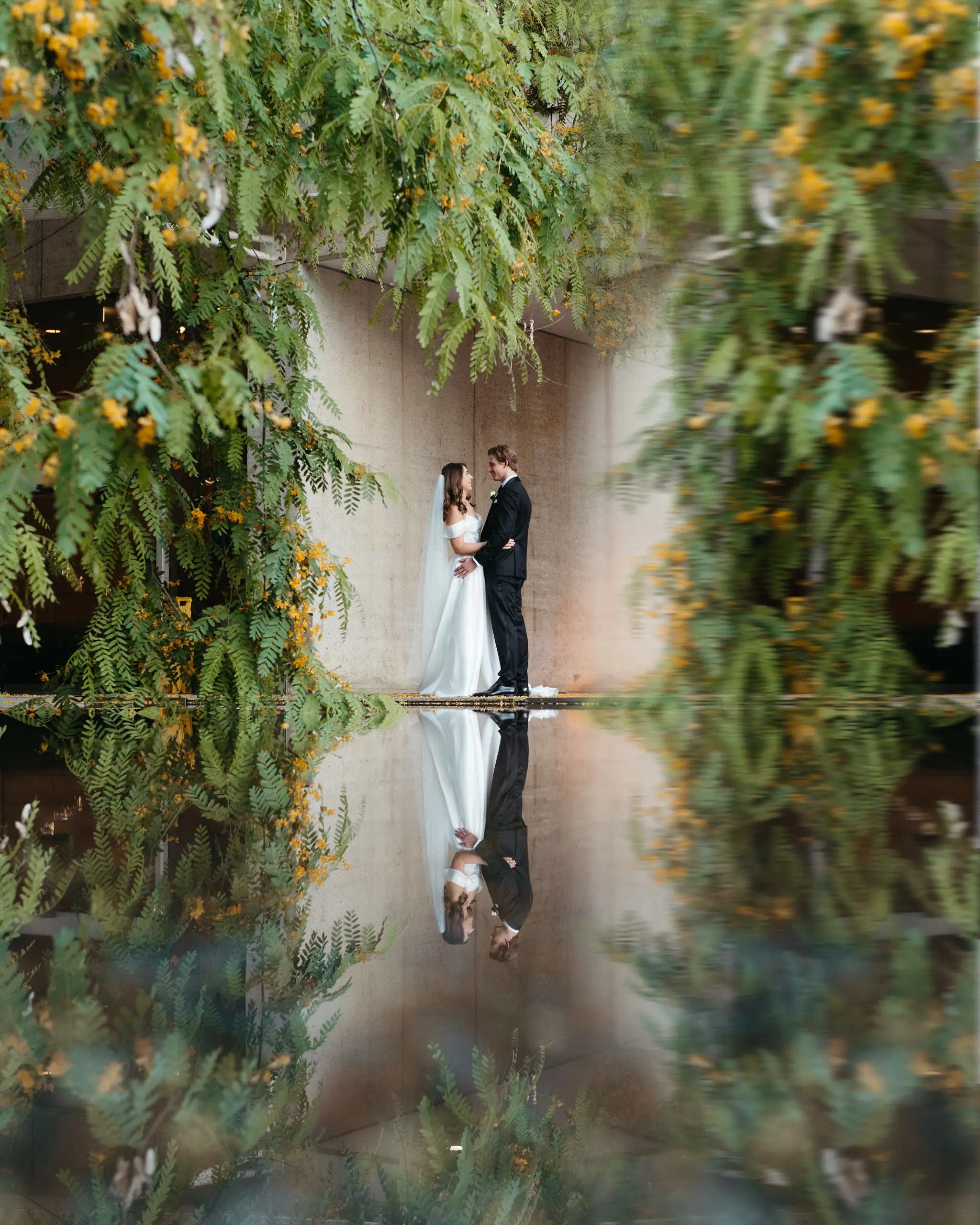 Bride and groom beneath an arch of hanging blooms, mirrored in the pool below