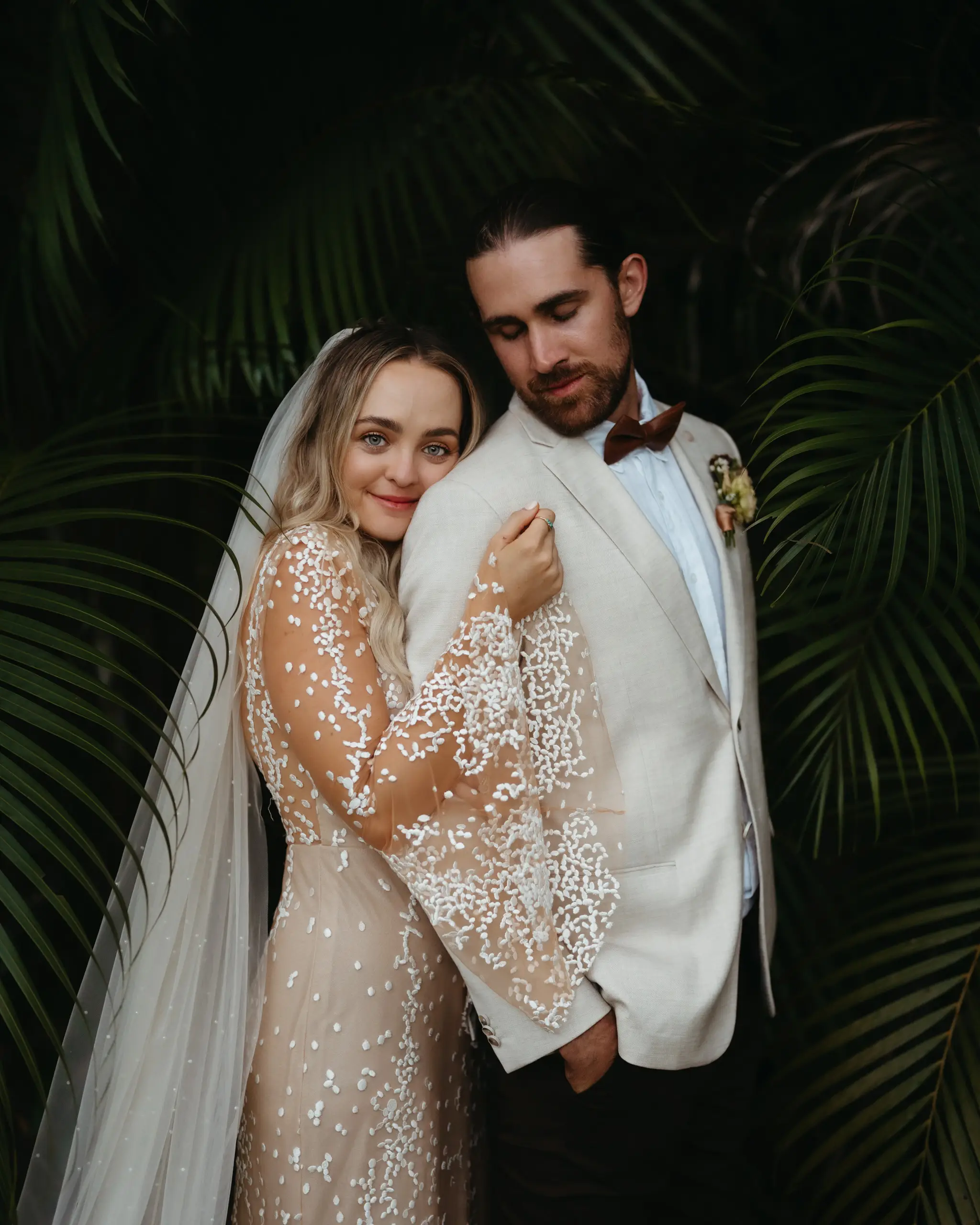 Bride leaning into the groom's white jacket beneath the palms