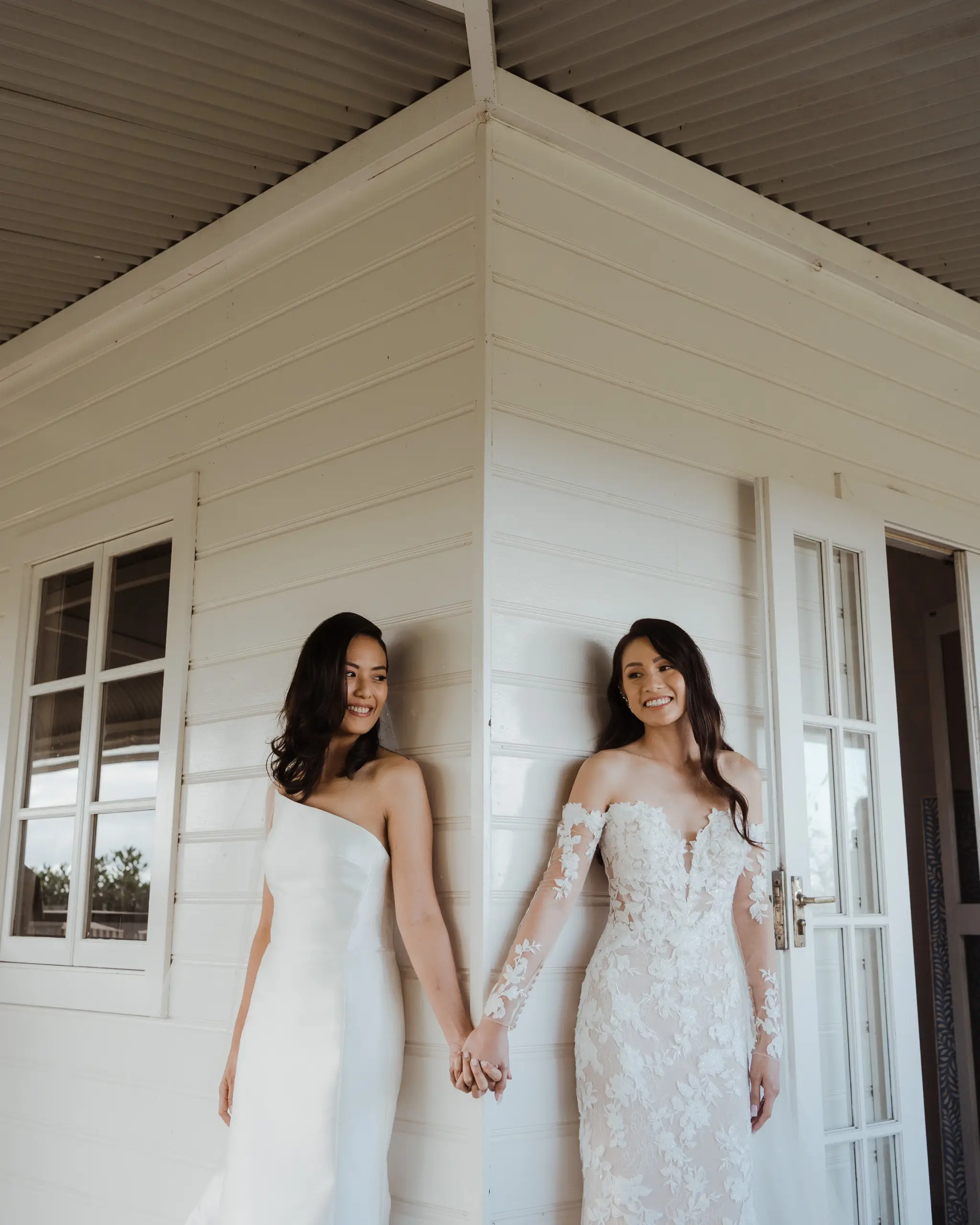 Two brides holding hands on the veranda of a white cottage