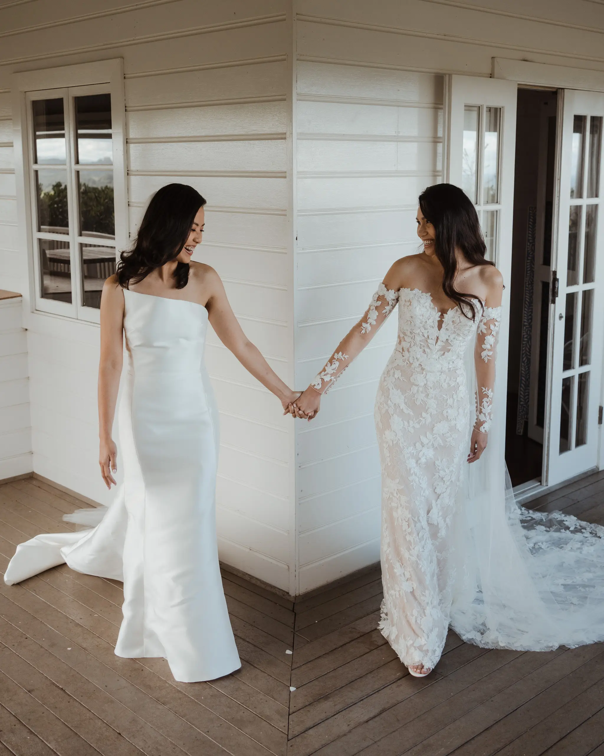 Two brides hand in hand at the corner of a white weatherboard cottage