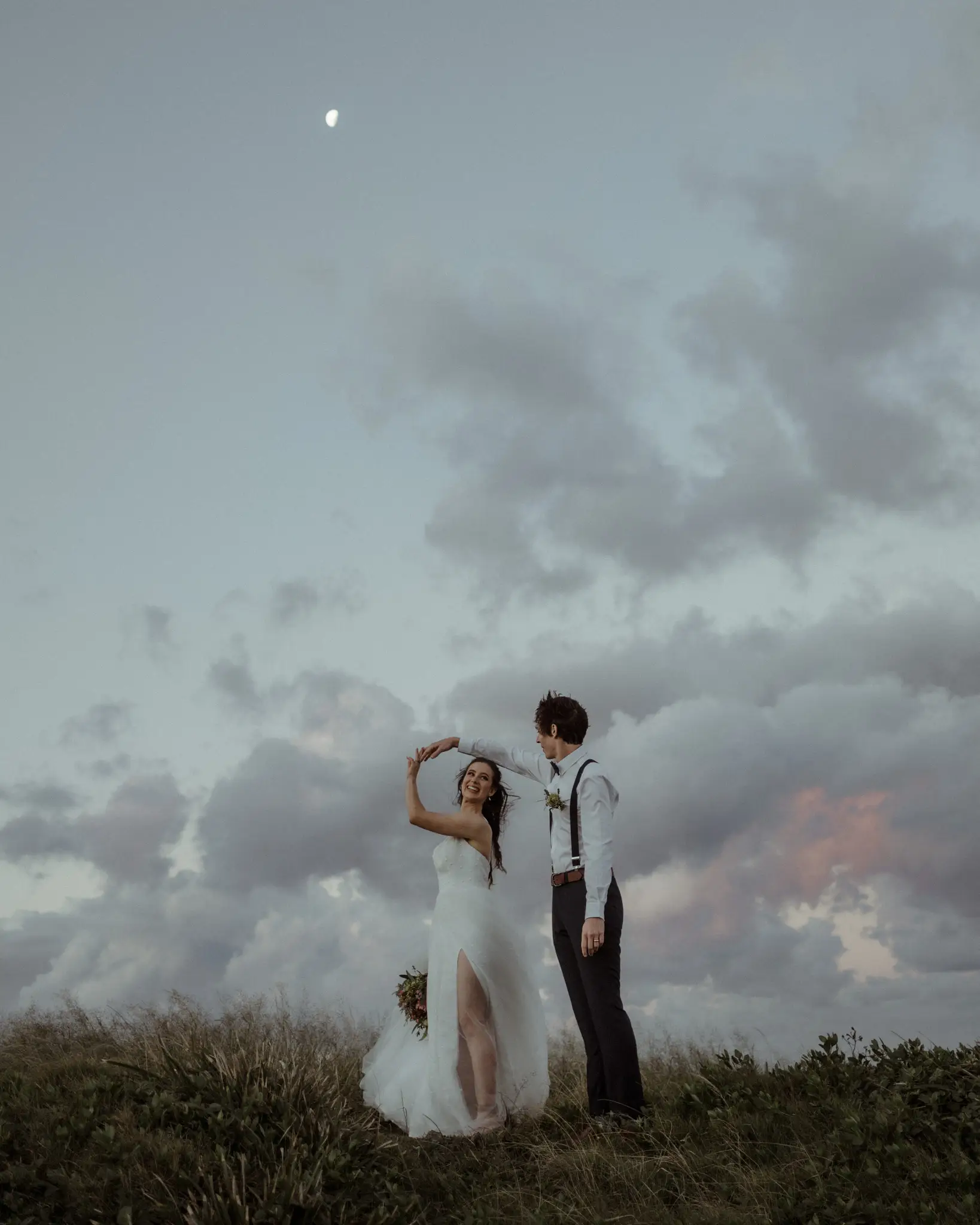 Bride raising her flower crown on a headland under a moody sky