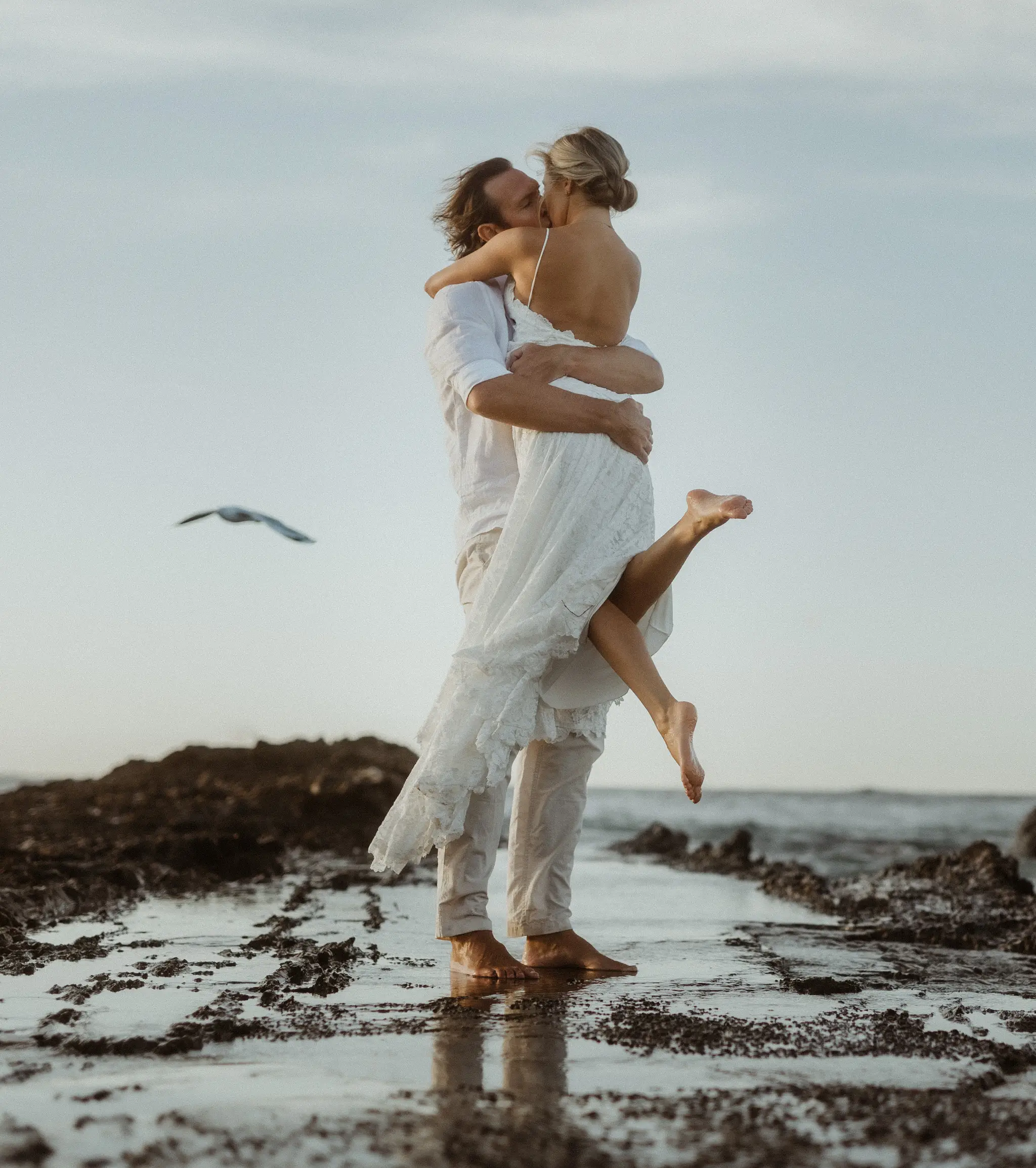 Groom lifting the bride barefoot on the rock shelf, a gull passing