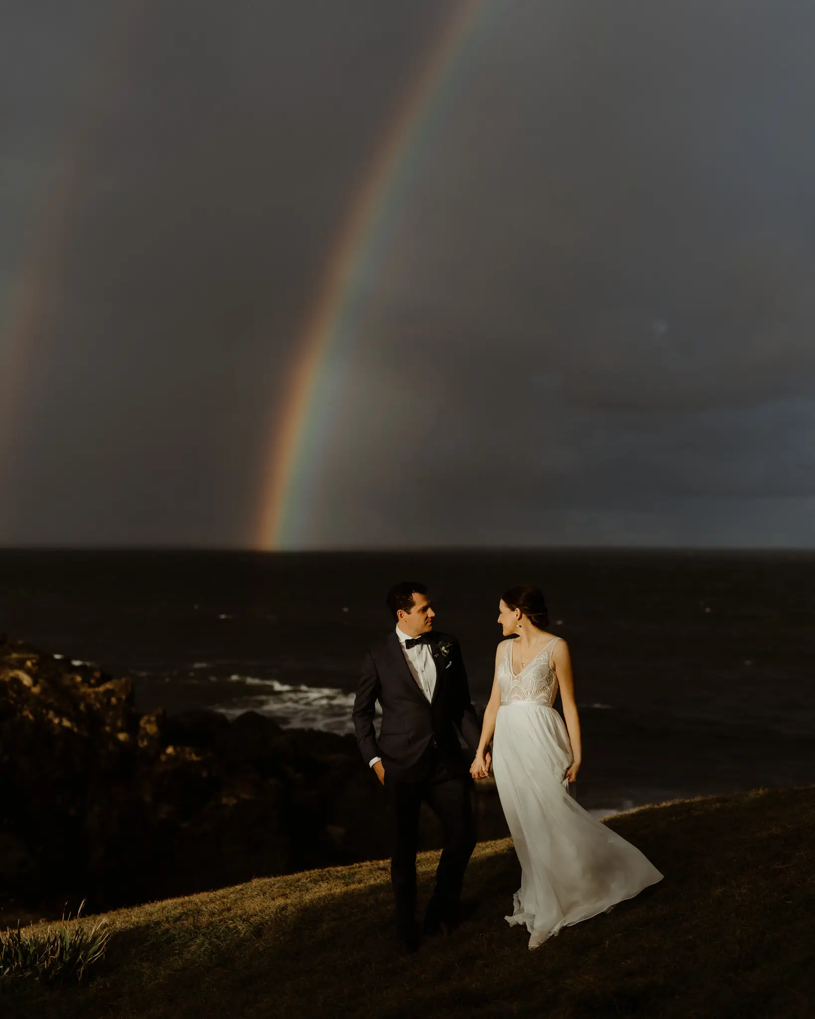 A rainbow landing on the sea behind the couple on the cliff edge