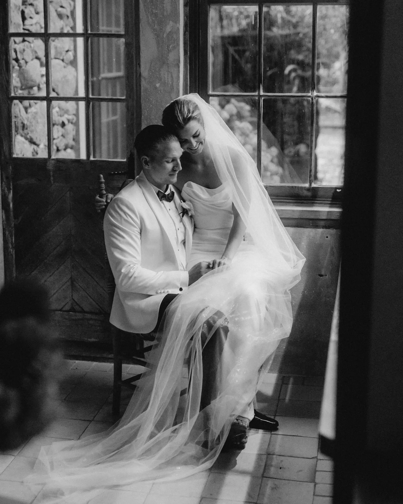 Couple seated in tall window light, veil across their laps, in black and white