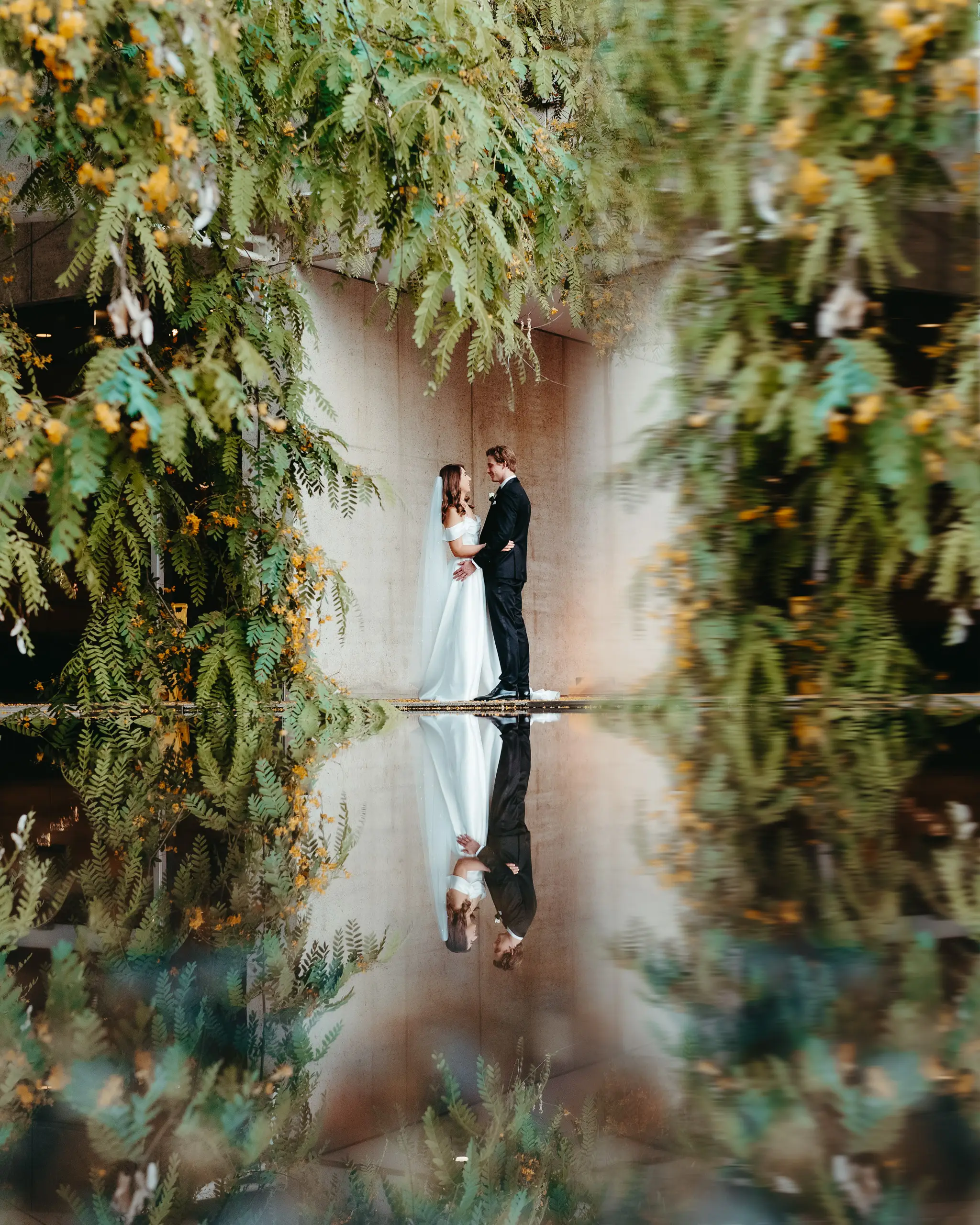 Bride and groom under hanging blooms, mirrored in the reflection pool
