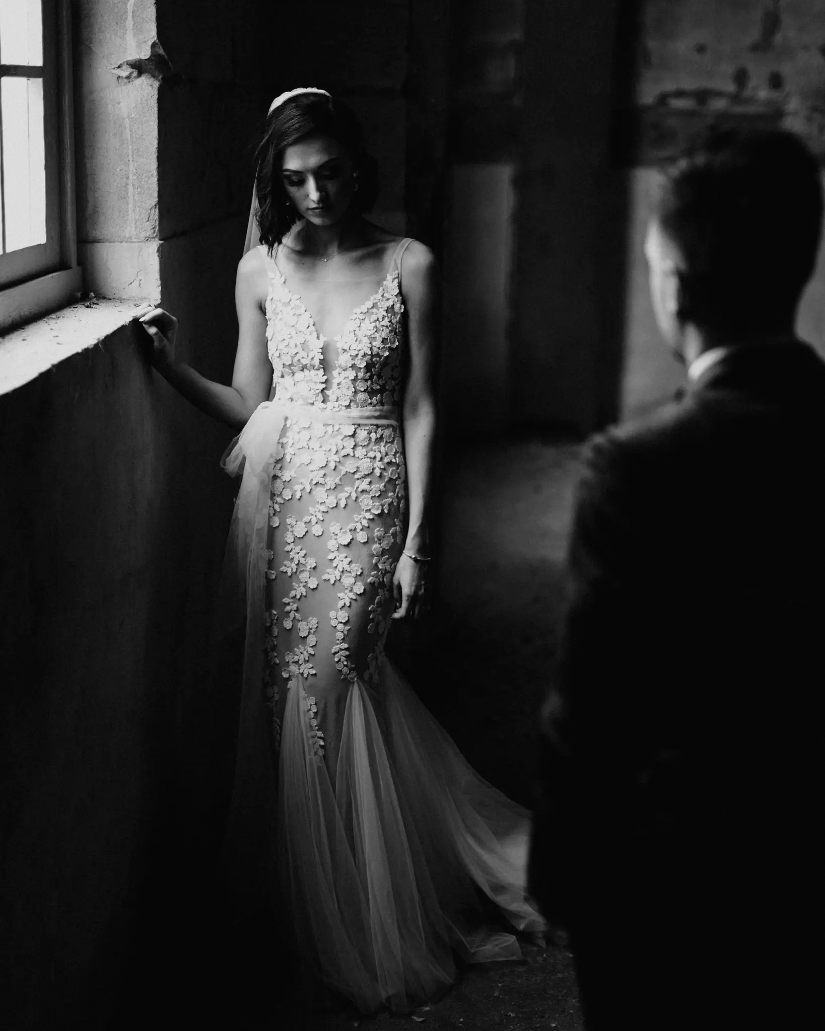 Bride by a stone window, groom watching