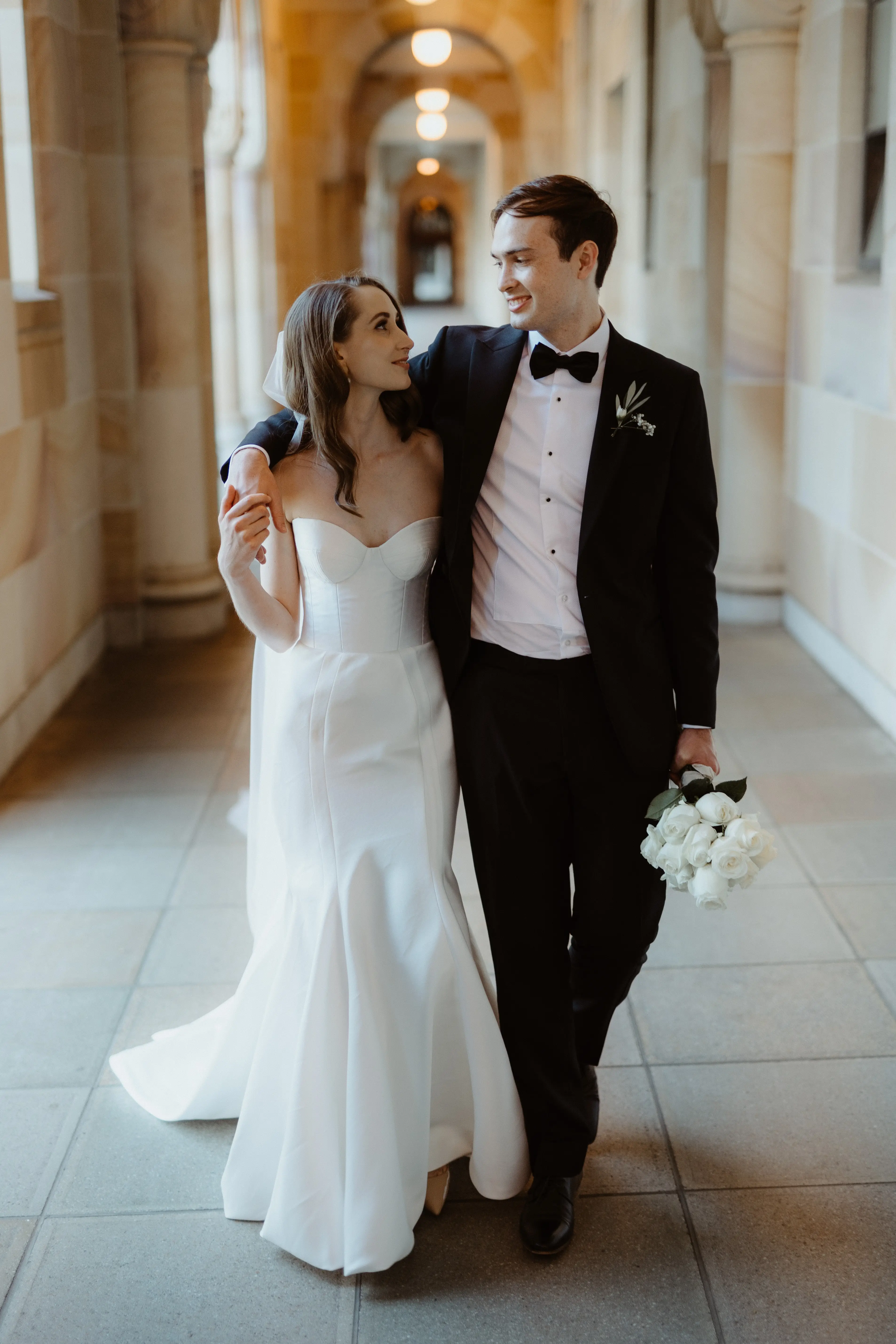 The couple arm in arm down a warm sandstone colonnade, Brisbane