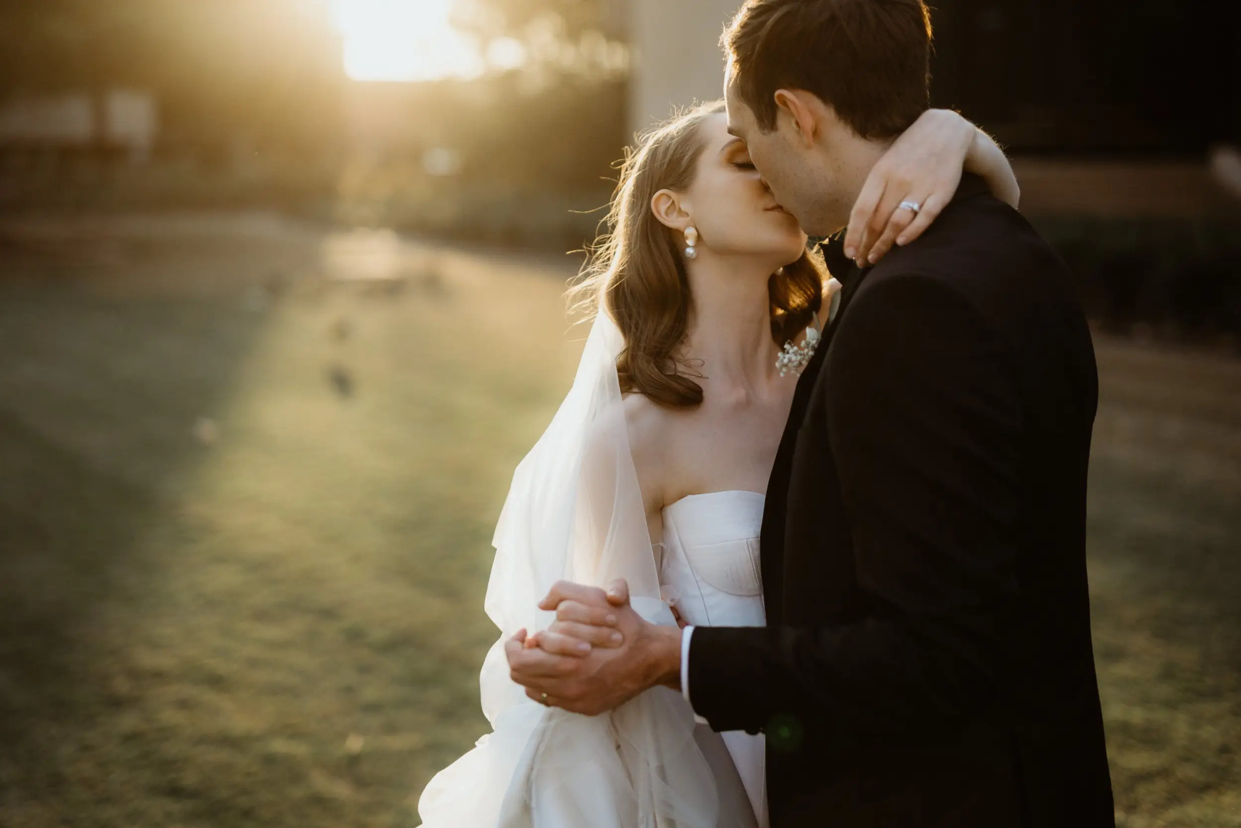 A couple kissing on the lawn in low golden sunlight, her veil catching the light behind her