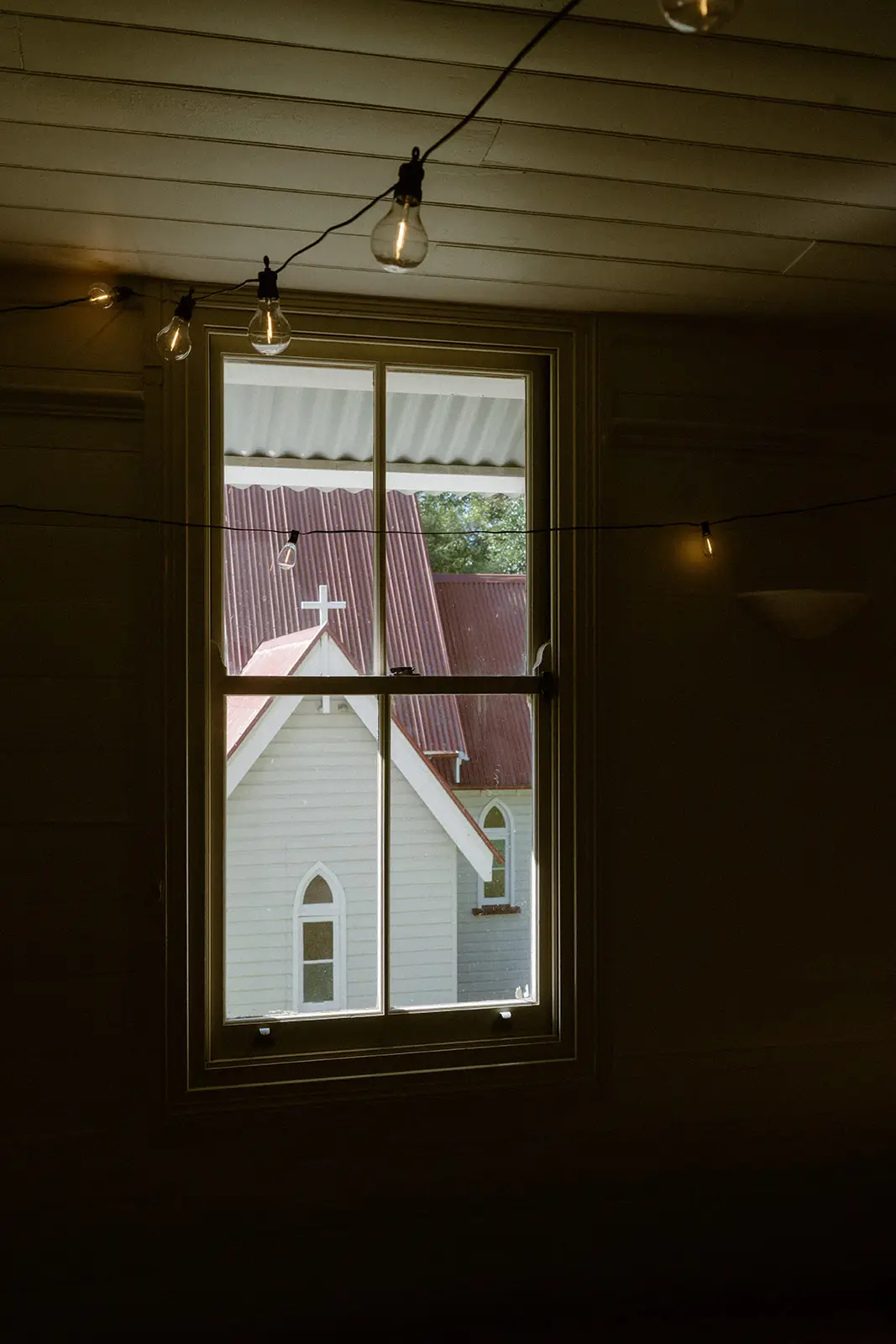 The church seen through a hall window, festoon bulbs strung across the frame, Ewingsdale Hall