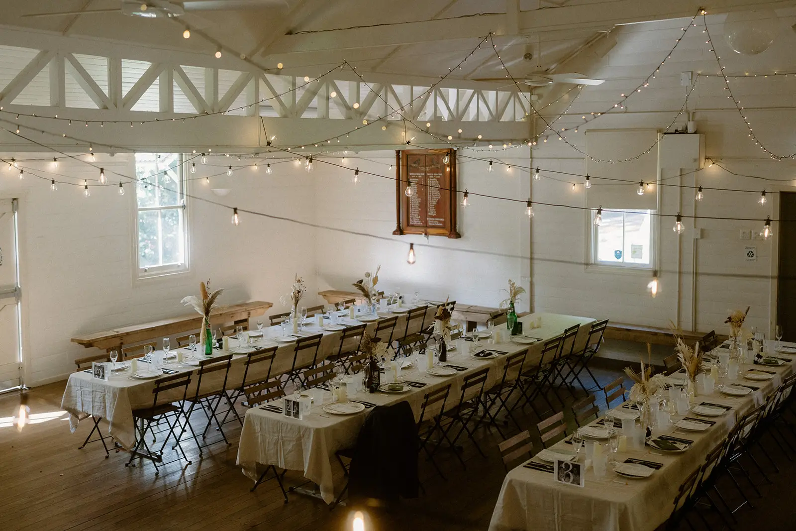 Long tables set the length of the hall under festoon lights and exposed white trusses, Ewingsdale Hall, Byron Bay