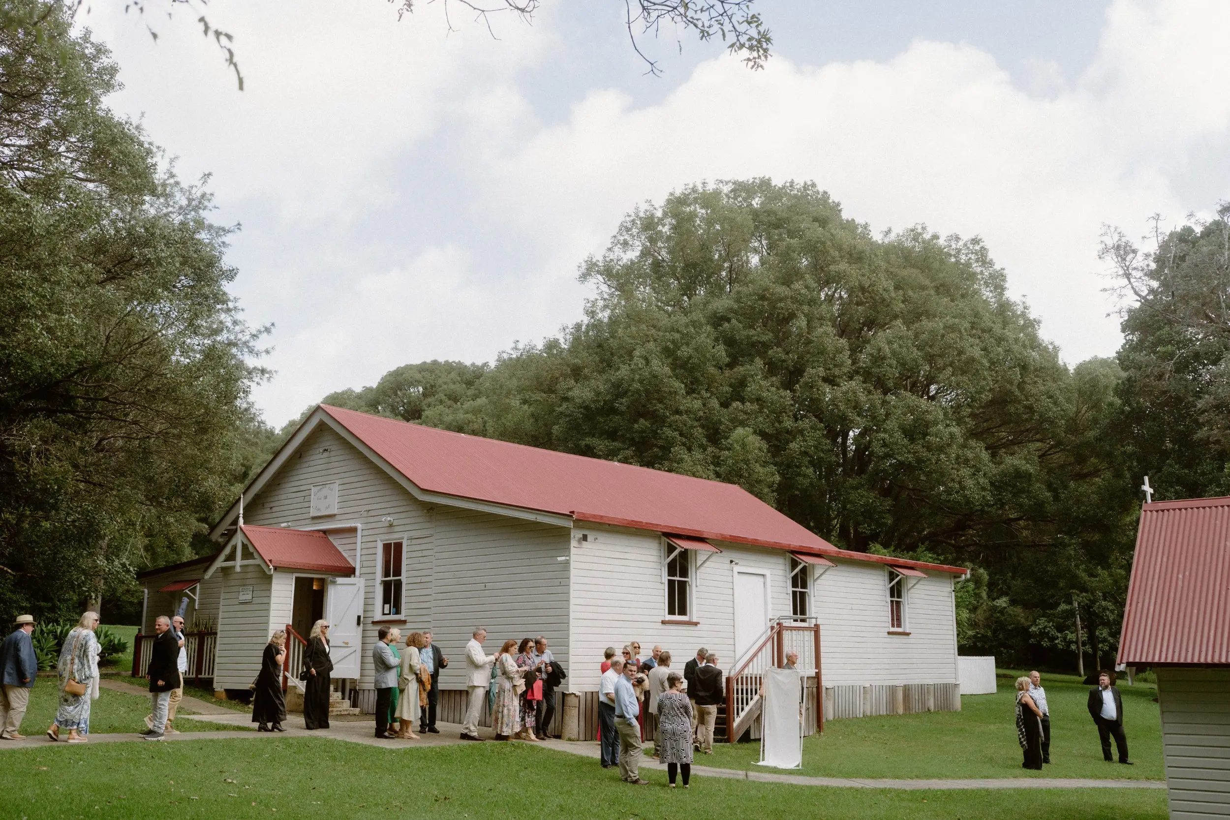 Guests gathered on the lawn outside the white timber hall with its red roof, the fig canopy behind, Ewingsdale Hall, Byron Bay