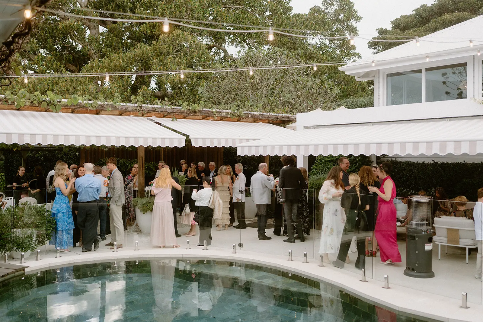 Guests with drinks around the pool under festoon lights, the farmhouse behind, Fig Tree Restaurant