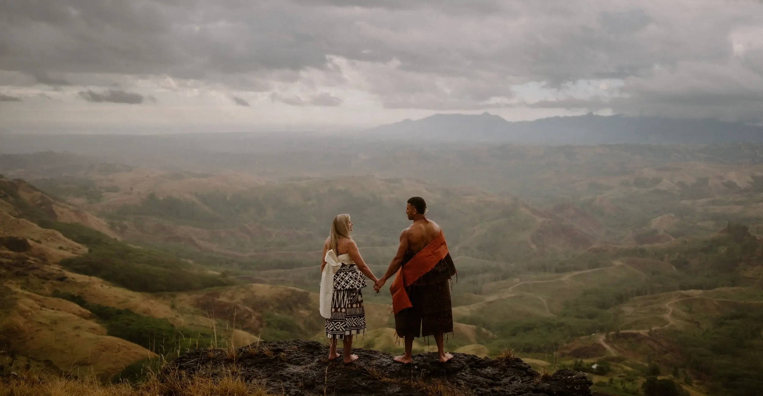 Brittany and Viliame holding hands on a rocky outcrop above the Fijian highlands under storm clouds, wearing traditional masi