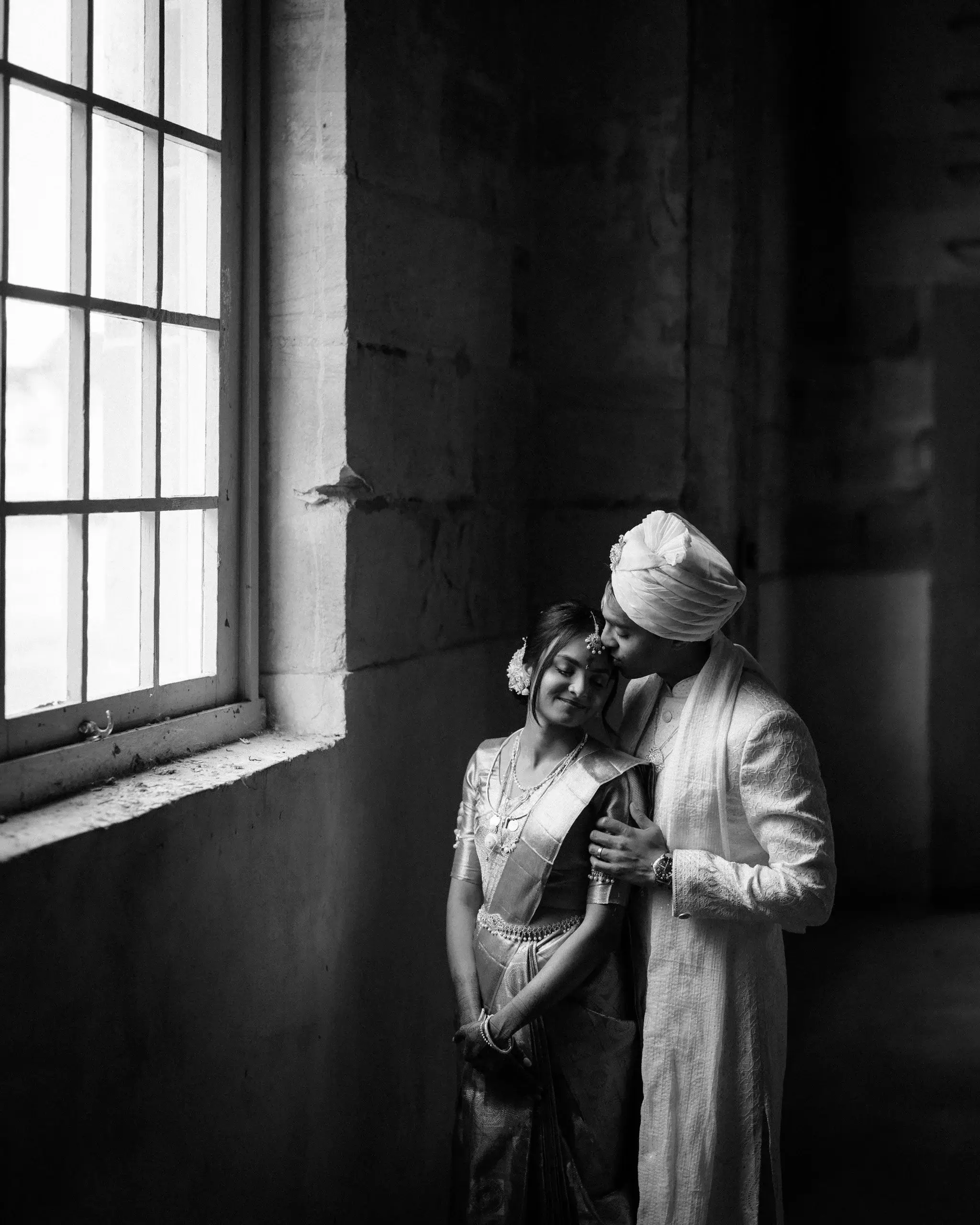 Black and white portrait of a couple in traditional dress beside a tall warehouse window