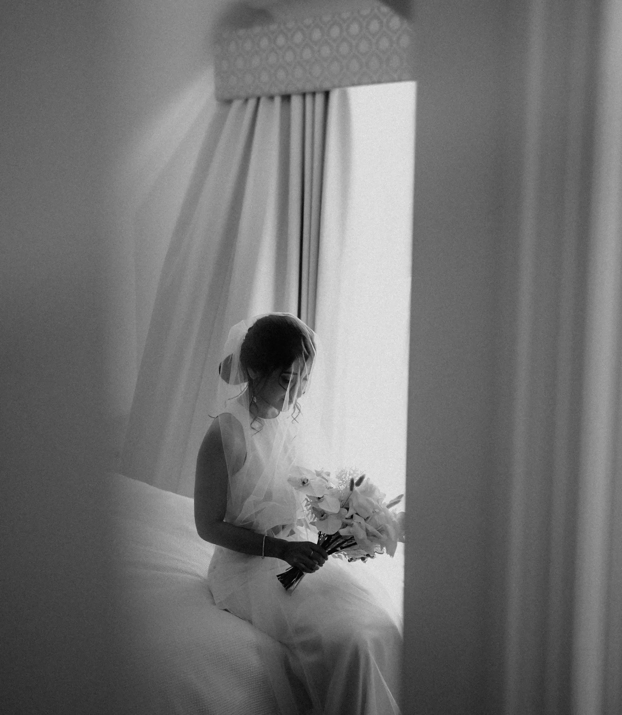 Black and white photograph of a bride seated by a window holding her bouquet