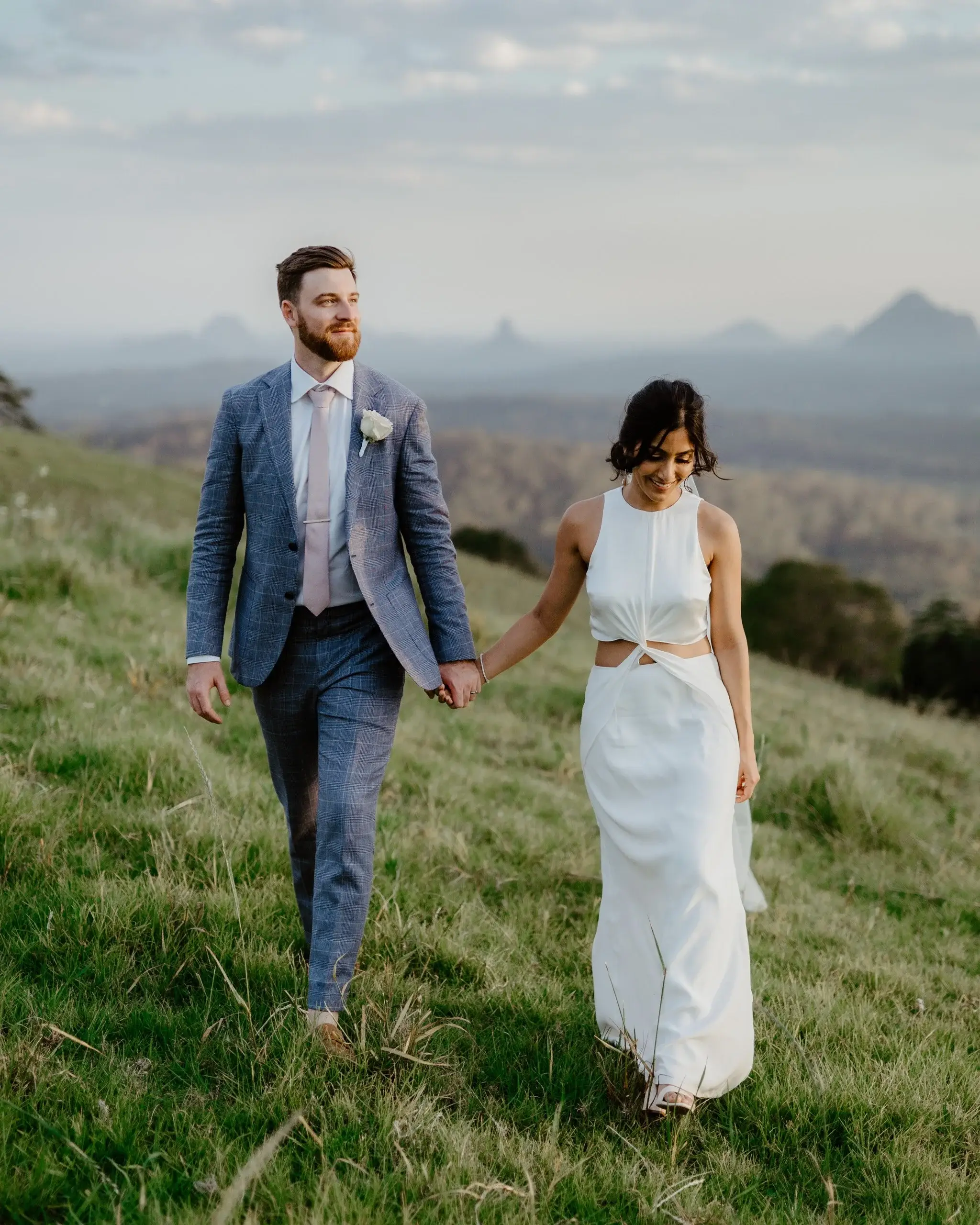 A couple walking a grassy ridge, the Glass House Mountains in the distance