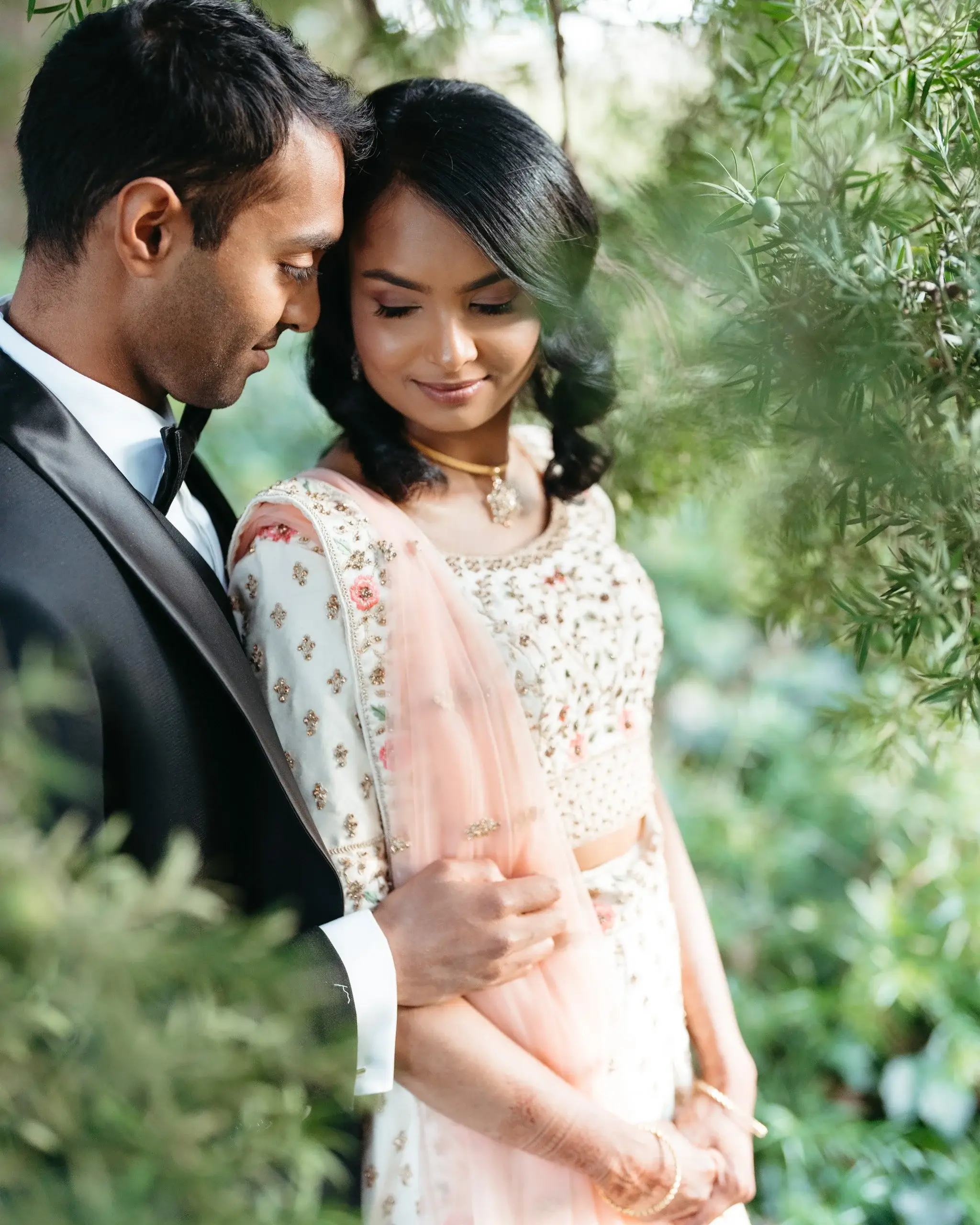 A couple standing together among olive-green foliage, the bride in an embroidered sari
