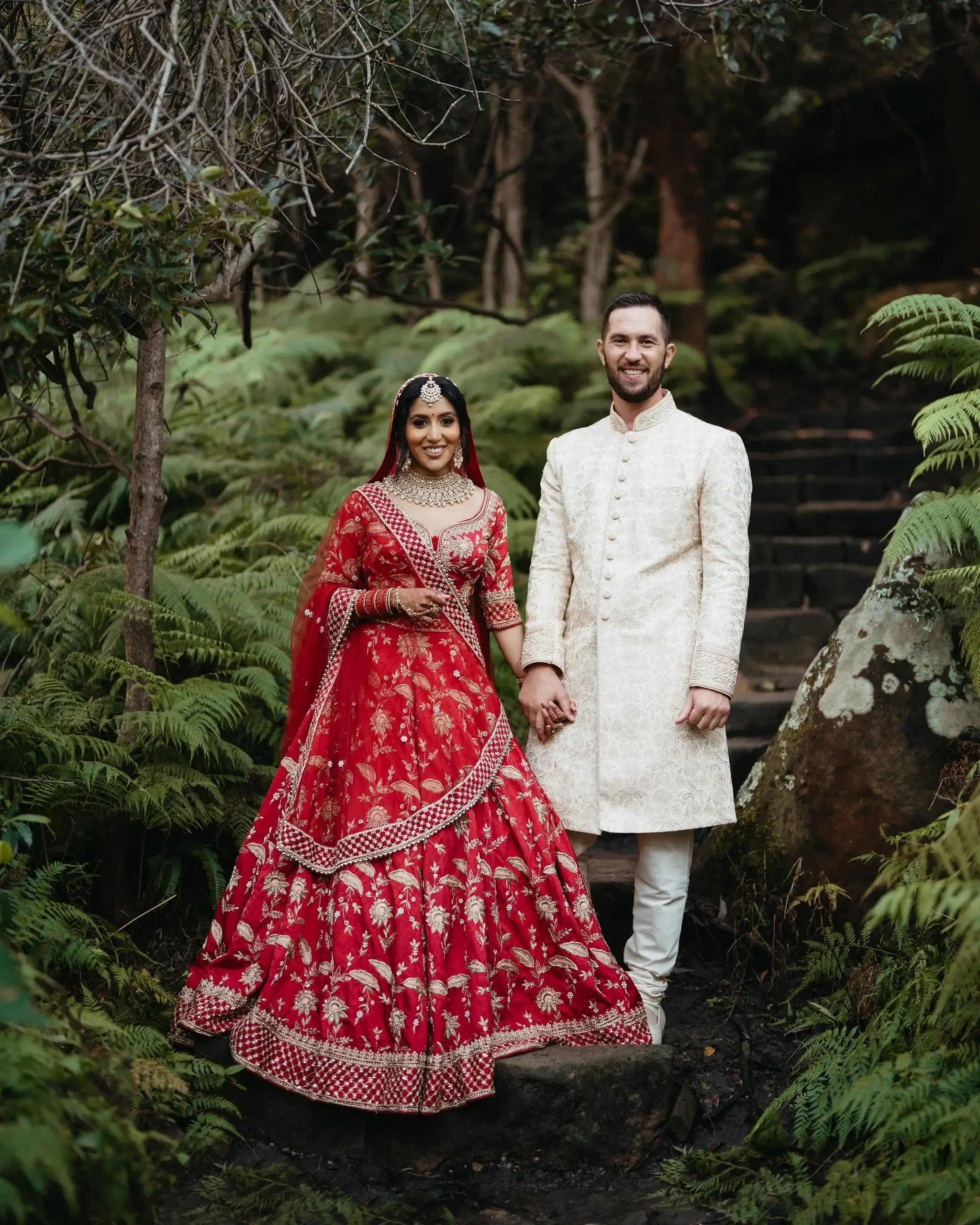 A couple in a red lehenga and cream sherwani standing in rainforest