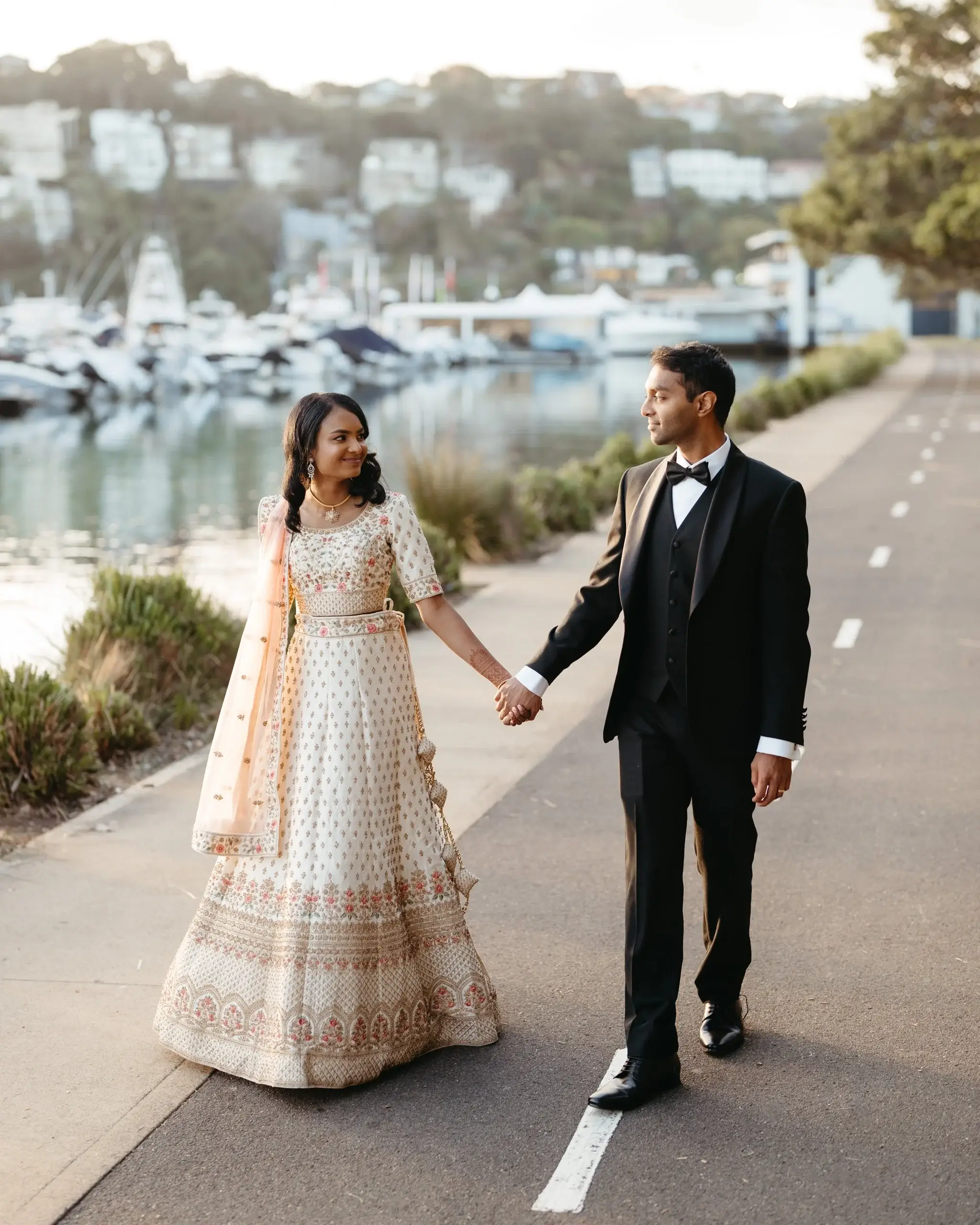 A couple holding hands on a road above a marina, the bride in a gold lehenga