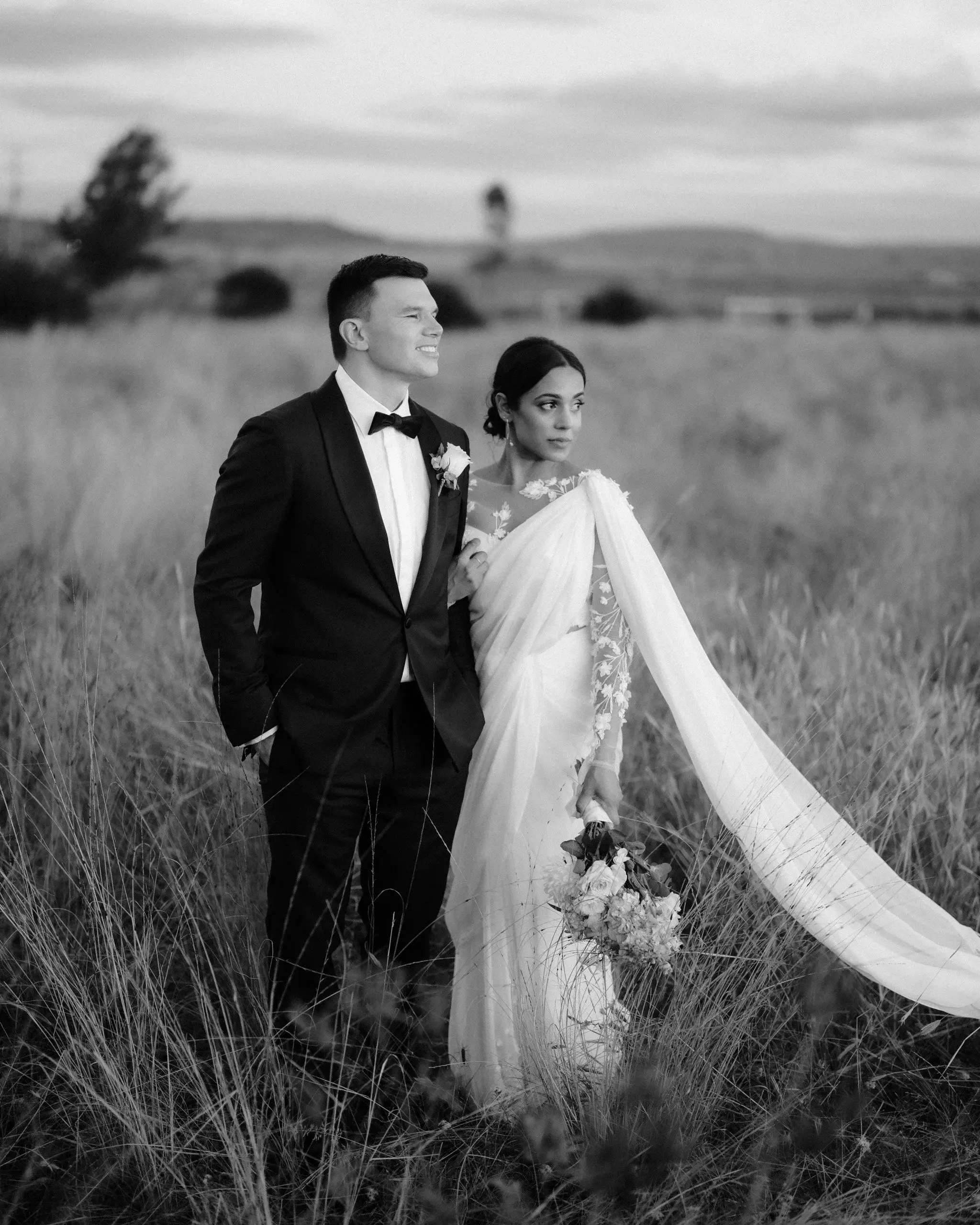 Black and white photograph of a couple standing in long grass, the bride’s veil trailing