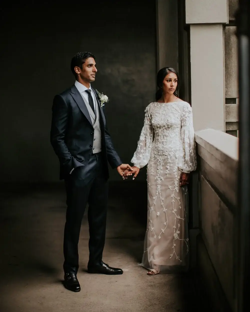 A couple holding hands in a bare concrete corridor, the groom in a navy three-piece suit and the bride in a white embroidered gown