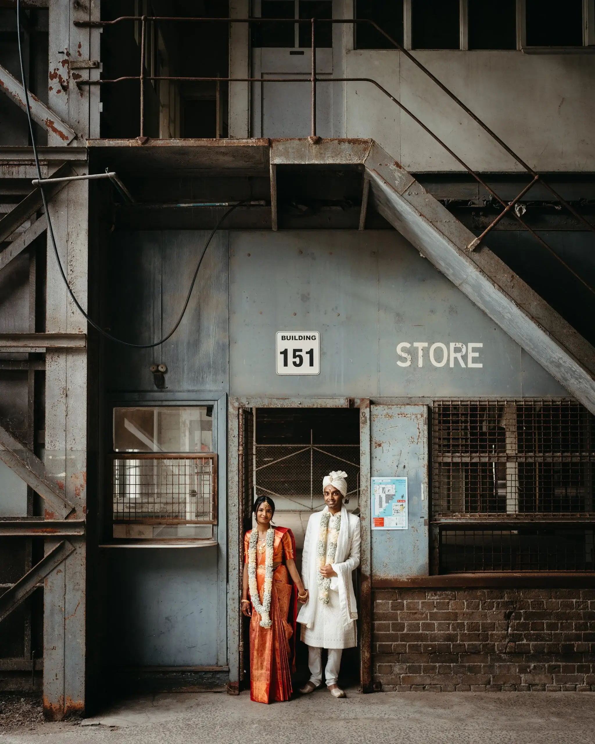 A bride in a red and orange sari and a groom in a white sherwani and turban standing in the doorway of a disused industrial building