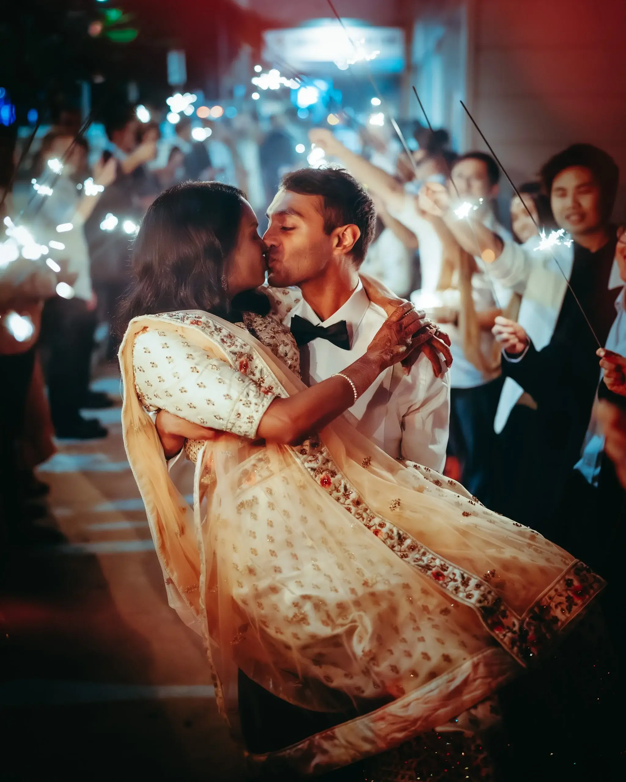 A couple kissing in the middle of a sparkler send-off, the bride in a cream and gold embroidered lehenga, guests holding sparklers along both sides