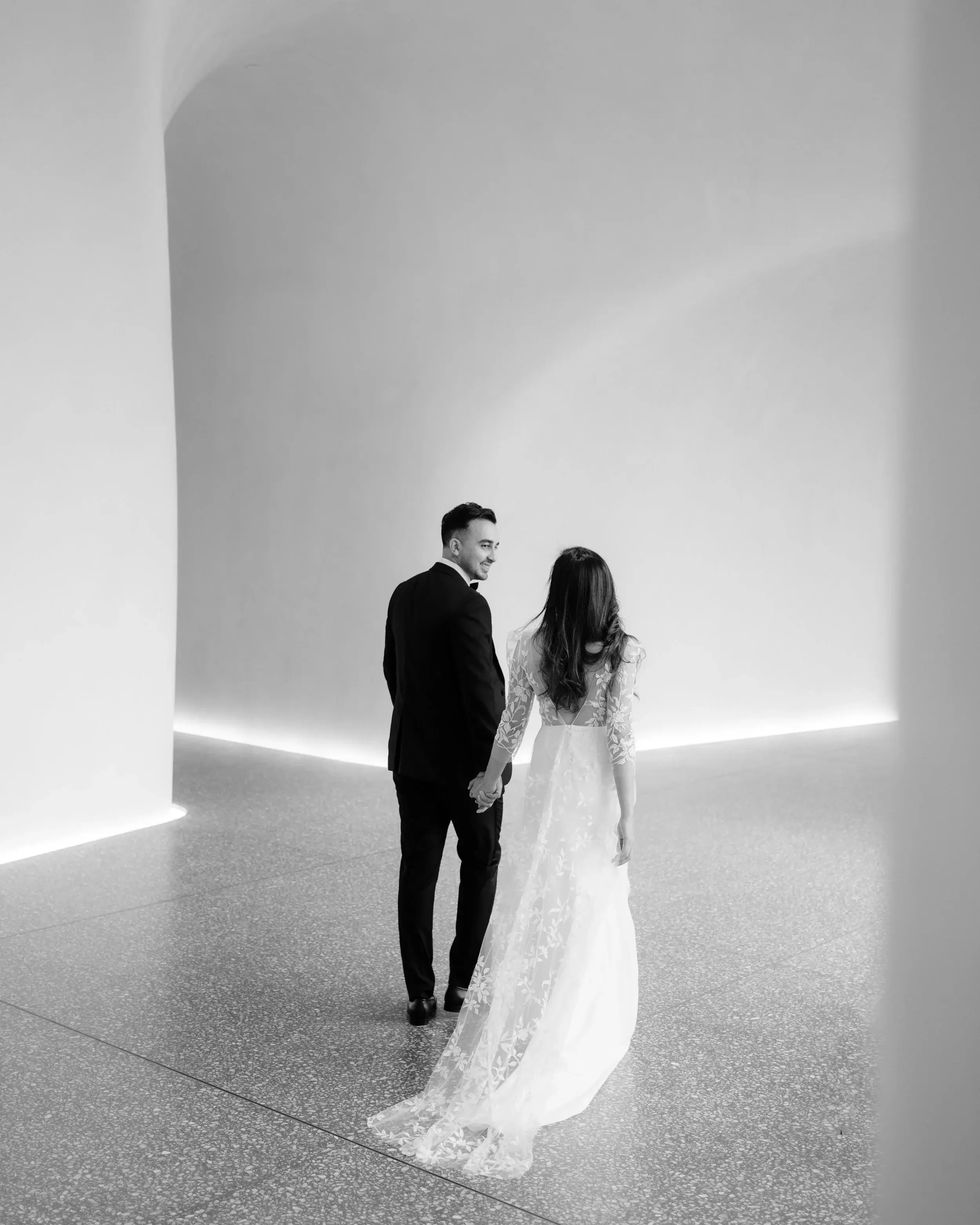 Black and white photograph of a couple standing in a vast curved white room, the bride's train trailing across the floor