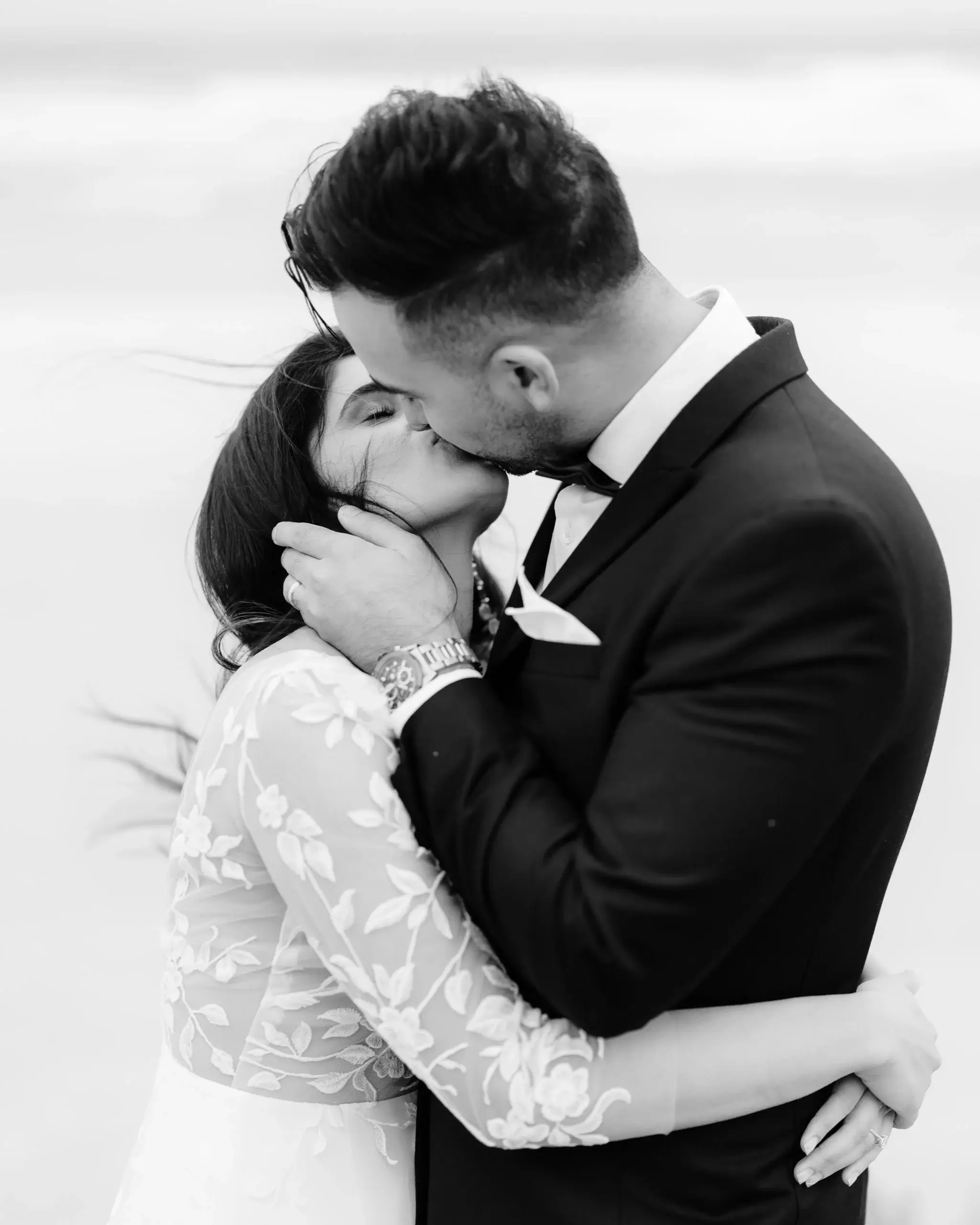 Black and white photograph of a couple kissing in the wind, the groom's hand at the bride's jaw