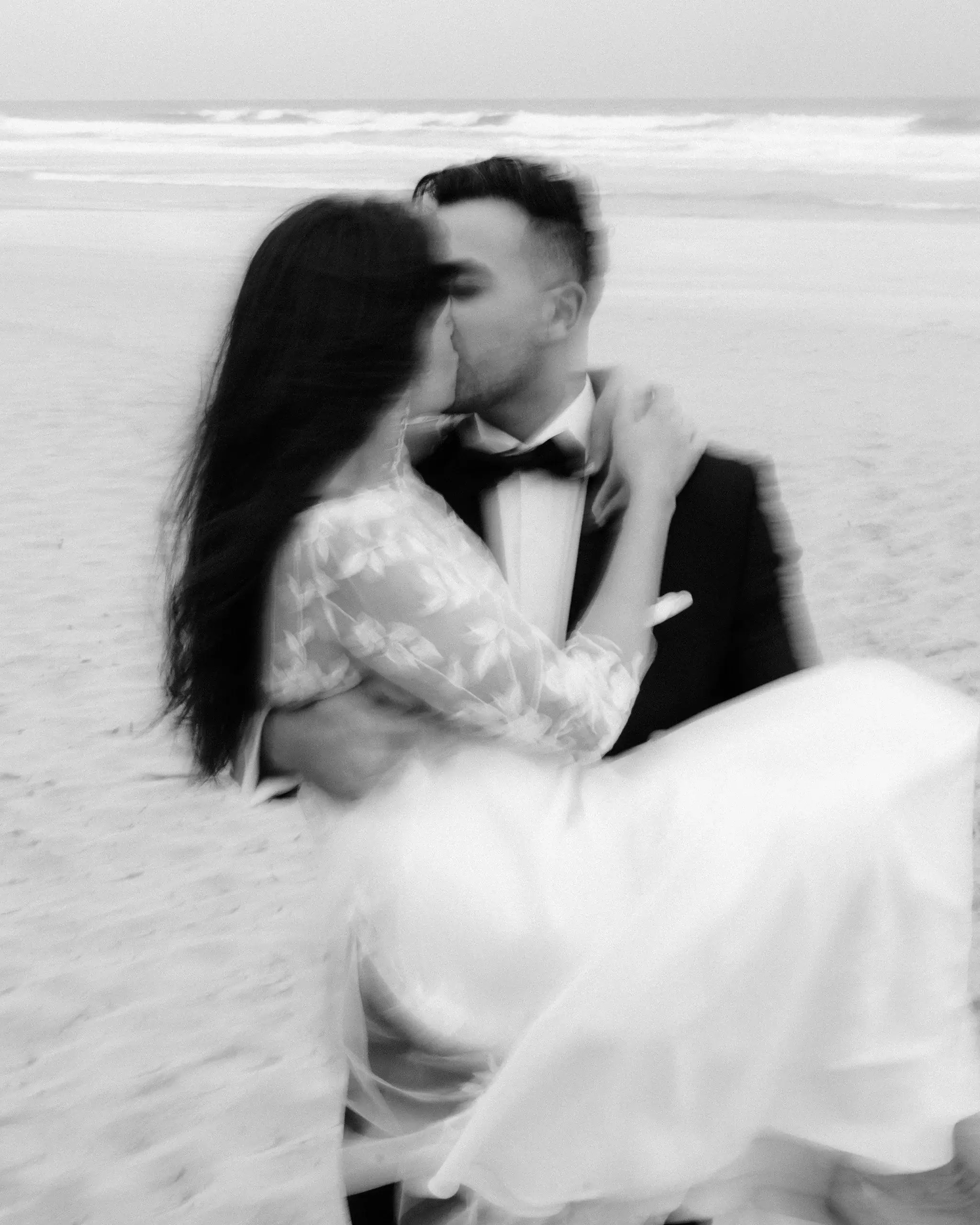 Motion-blurred black and white photograph of a couple kissing on the sand with surf behind them