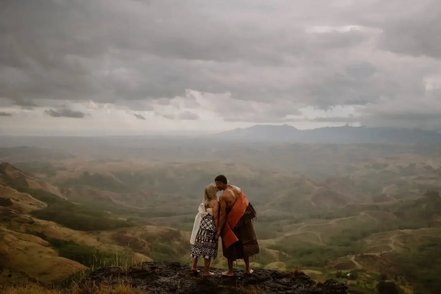 A couple kissing on a clifftop high above the Fijian highlands, wrapped in traditional masi cloth under heavy storm cloud