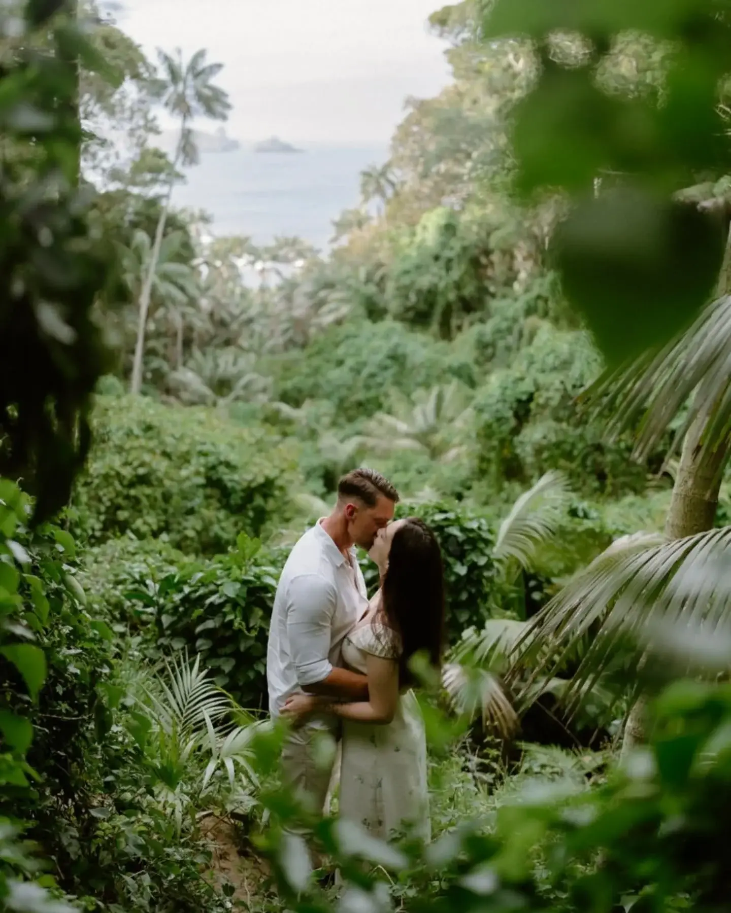A couple kissing among palms and dense green, the ocean showing through a gap in the canopy behind them