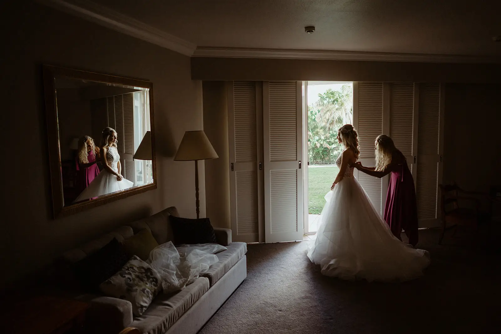 A bride having her gown fastened at the open doors of a suite, her reflection in the mirror behind, InterContinental Sanctuary Cove