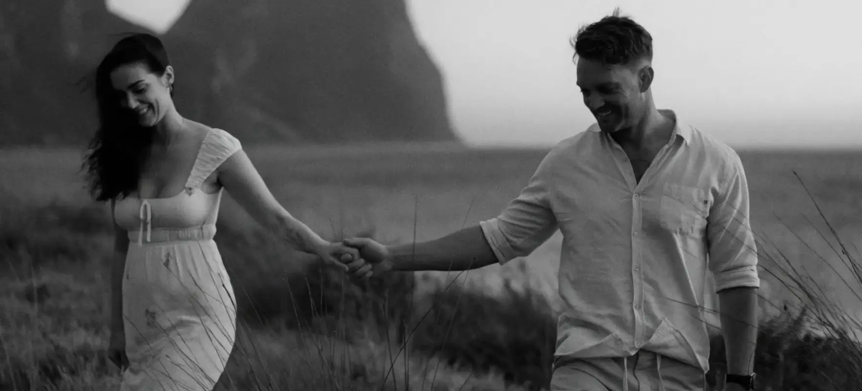 A couple walking hand in hand through long grass, both smiling, a headland rising through the haze behind them