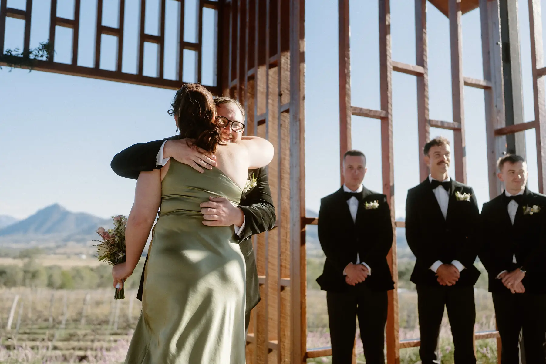 A hug with a guest in green satin, and Ryan waiting at the front of the chapel with his groomsmen and the ranges behind, Kooroomba Vineyard and Lavender Farm