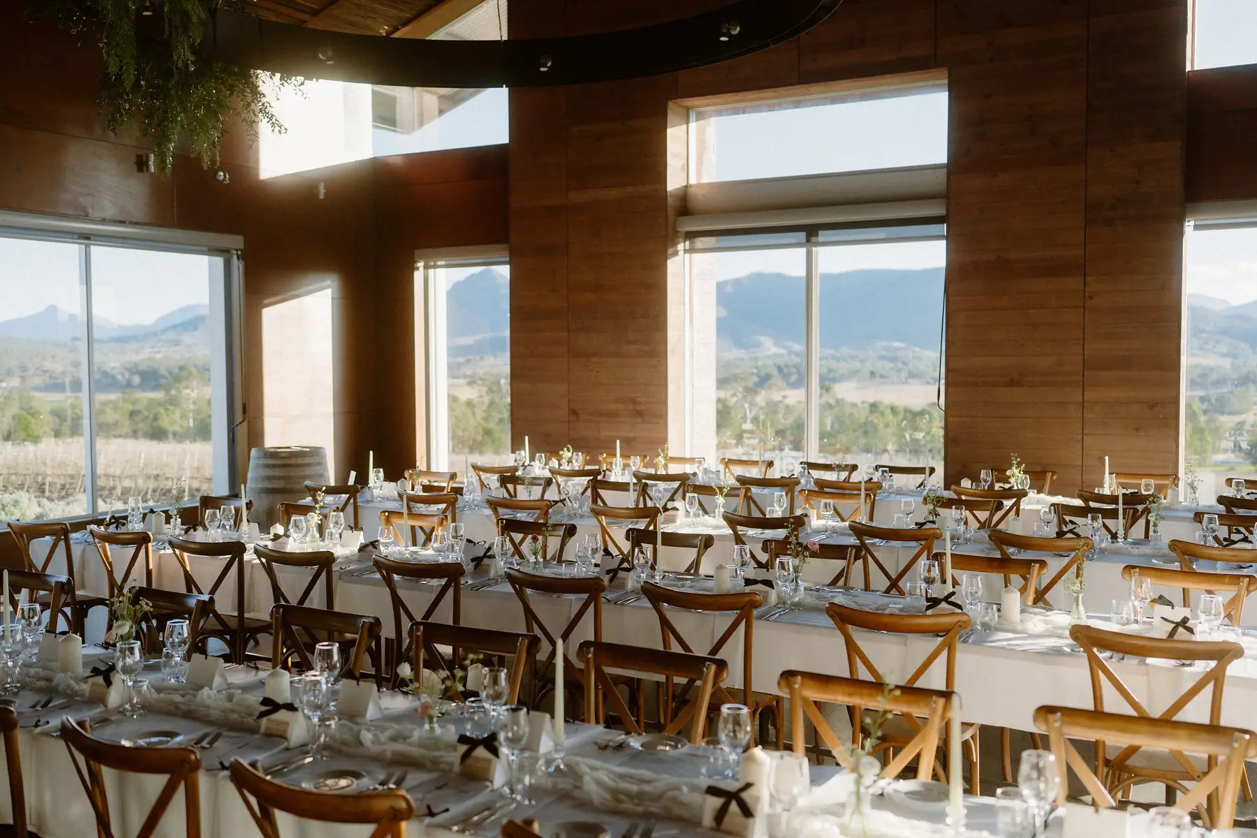 Long tables and cross-back chairs under the timber ceiling, windows open to the mountains, Kooroomba Vineyard and Lavender Farm