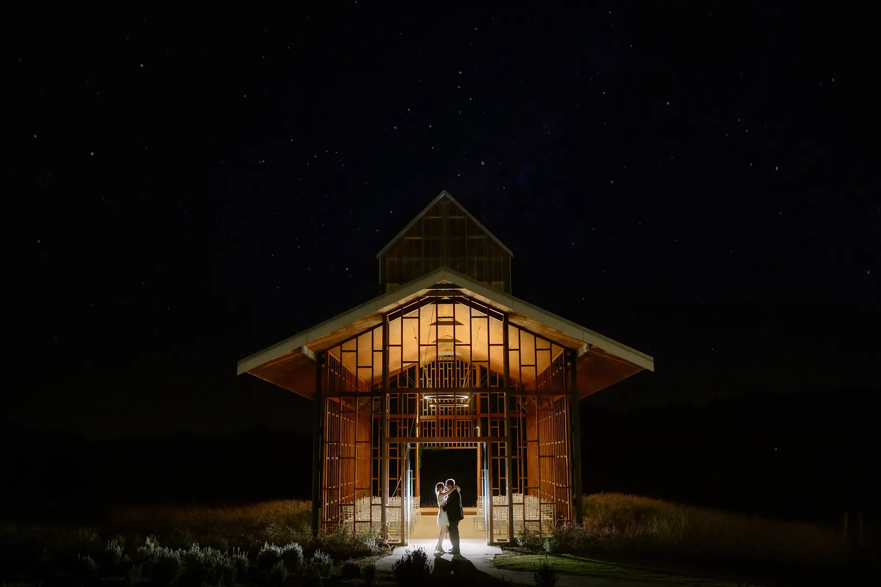 The chapel lit from within at night, two figures small in the doorway, Kooroomba