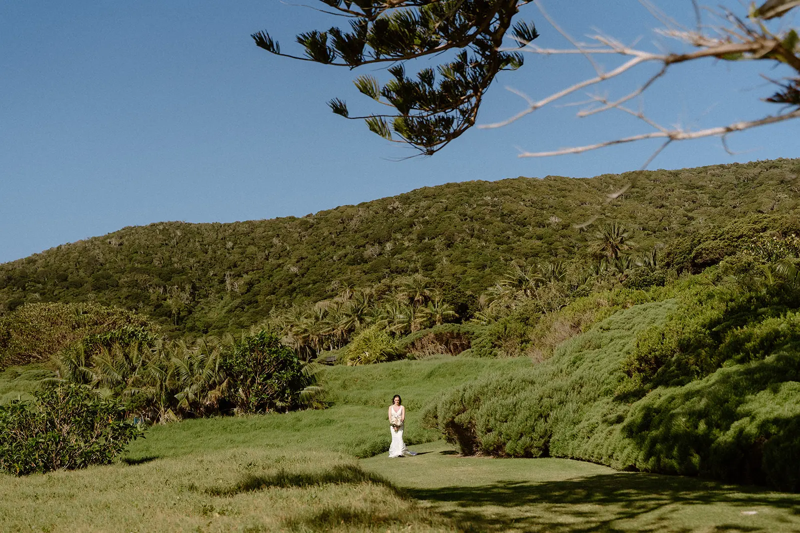 Mel walking across the lawn with her bouquet, forested hillside behind, Lord Howe Island