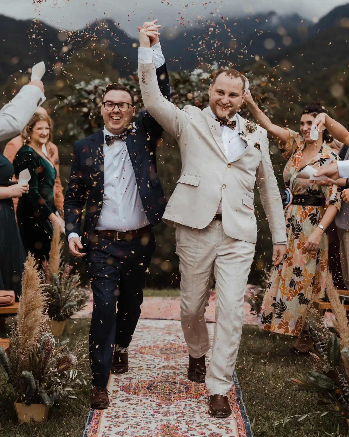 Two grooms walking back down the aisle hand in hand through a shower of petals, the ranges behind them, Mavis's Kitchen, Uki