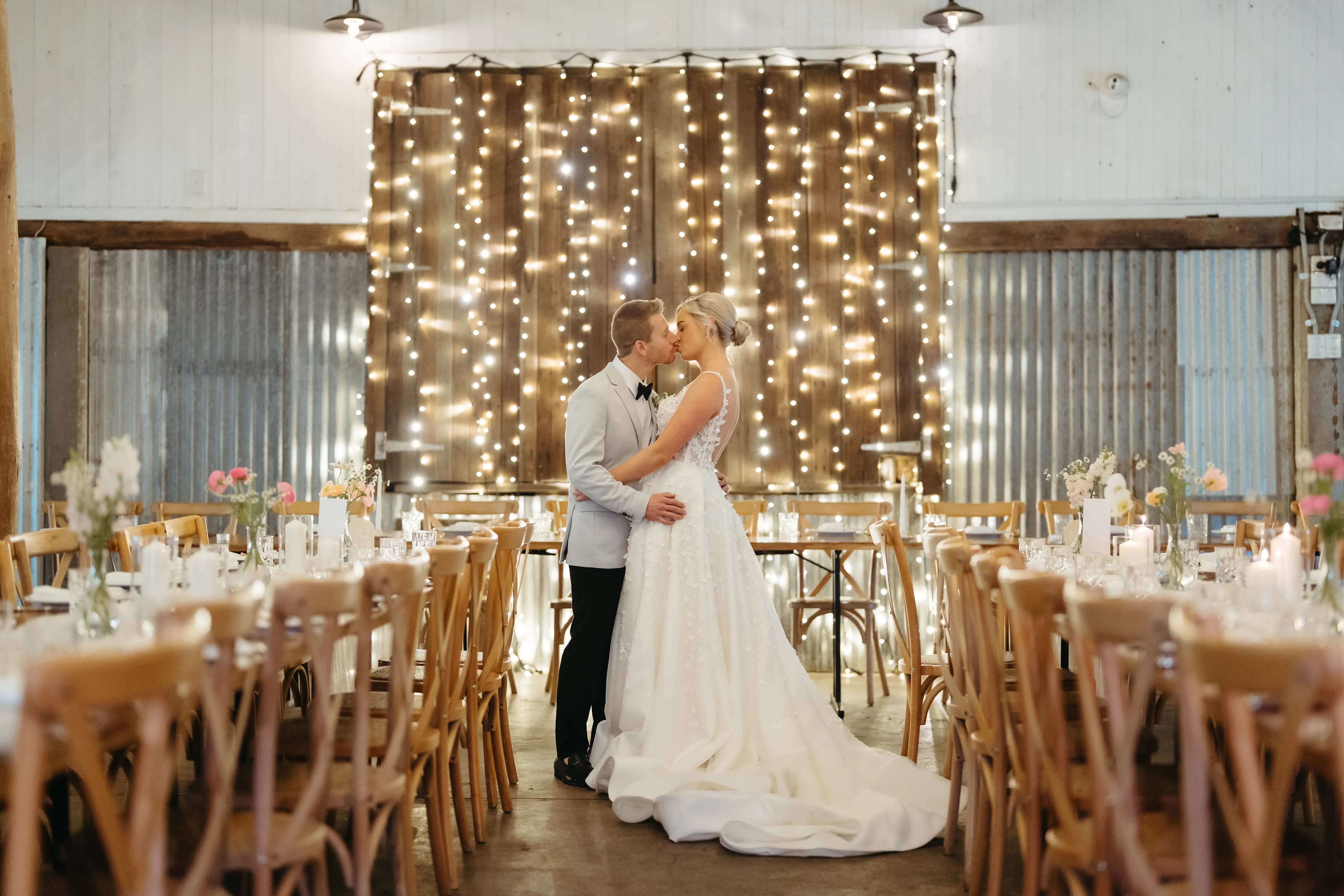 First look at the reception — the couple between the long timber tables and the fairy-light curtain in the 1902 barn, The Acre Boomerang Hills