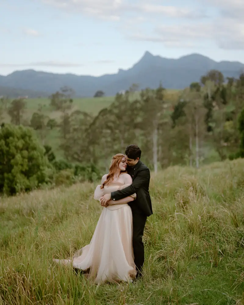 A couple holding each other in the long grass with Wollumbin and the Tweed ranges behind them, Sēlah Valley Estate, Limpinwood